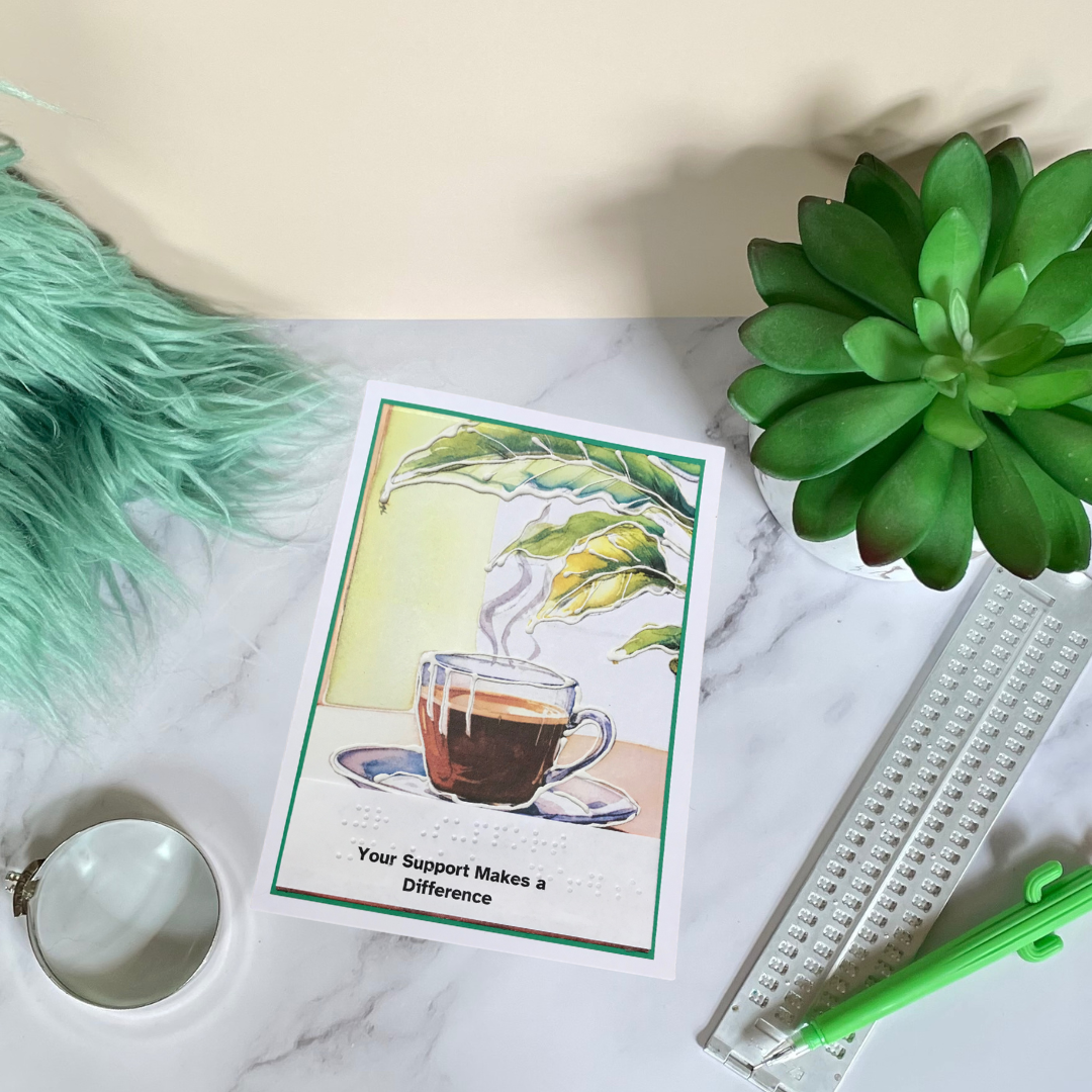 A handmade braille thank you card with an illustration of a steaming cup of coffee and a houseplant, placed on a marble surface with a green decorative element and a measuring tool in the background.