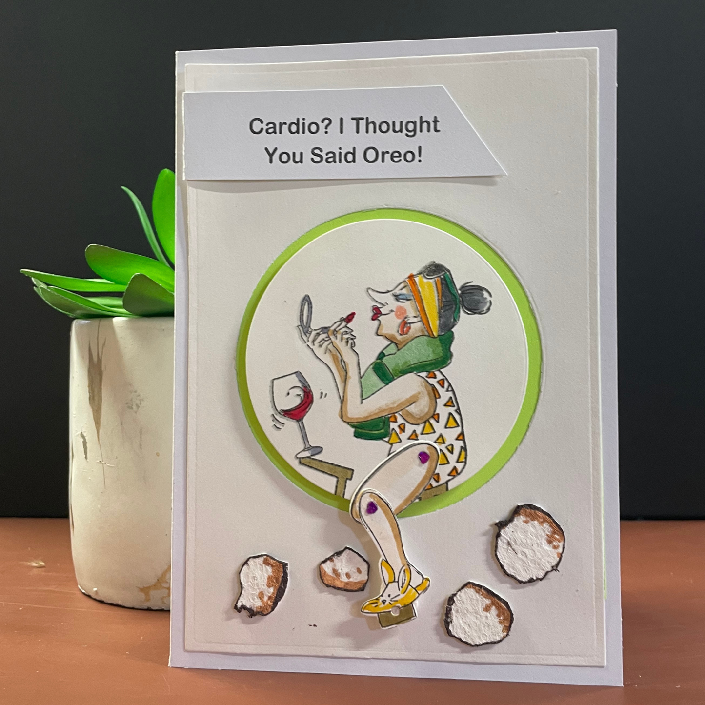 A handmade greeting card with a humorous message, featuring a 3D illustration of a woman on an exercise bike, a towel around her neck, and she is looking in a mirror adding lipstick, and oreo cookies on the floor. A green circular frame and the text 'Cardio? I Thought You Said Oreo!' at the top. The card is displayed on a wooden surface next to a small potted succulent against a dark gray background.