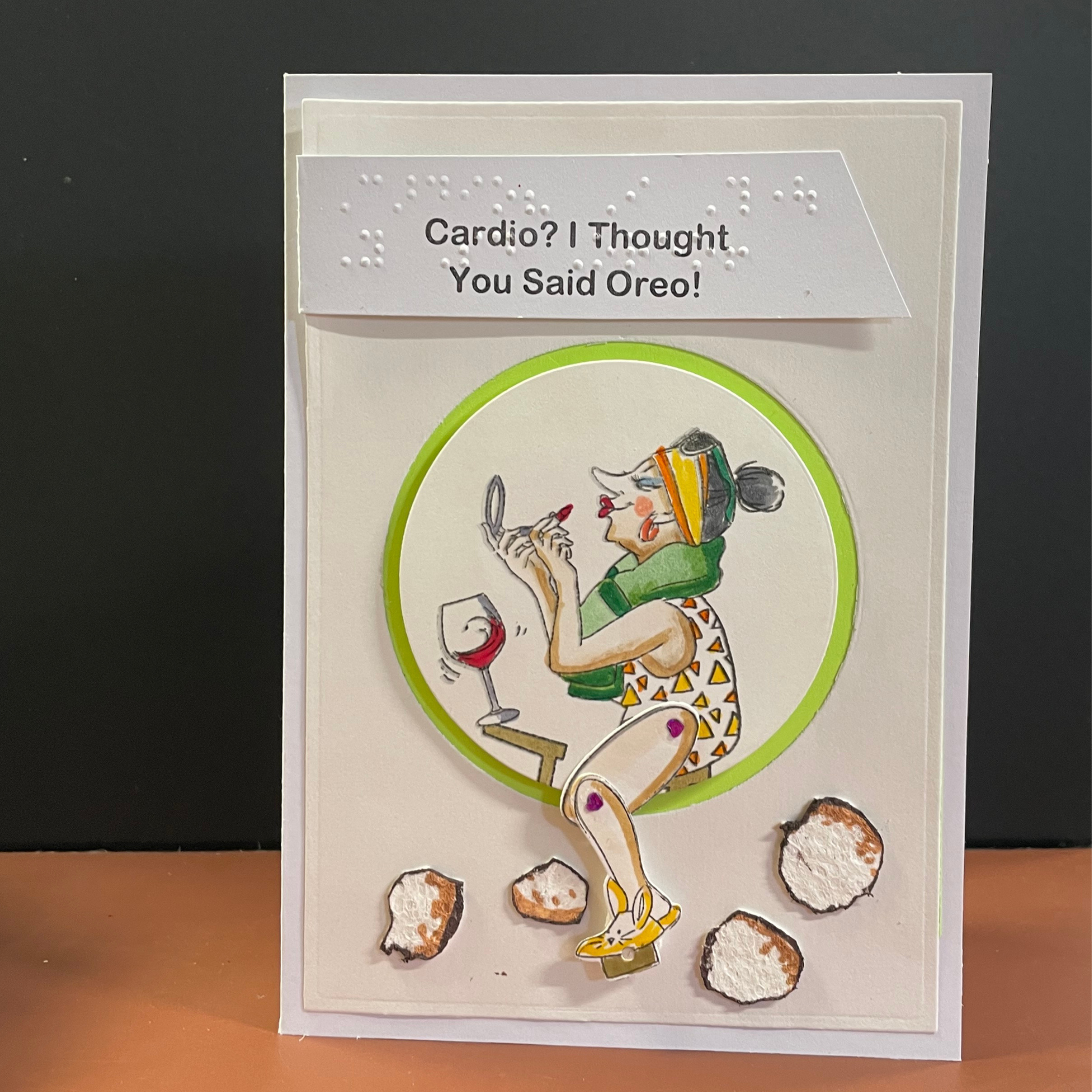 A handmade greeting card with a humorous message, featuring a 3D illustration of a woman on an exercise bike, a towel around her neck, and she is looking in a mirror adding lipstick, and oreo cookies on the floor. The text 'Cardio? I Thought You Said Oreo!' on a white header.