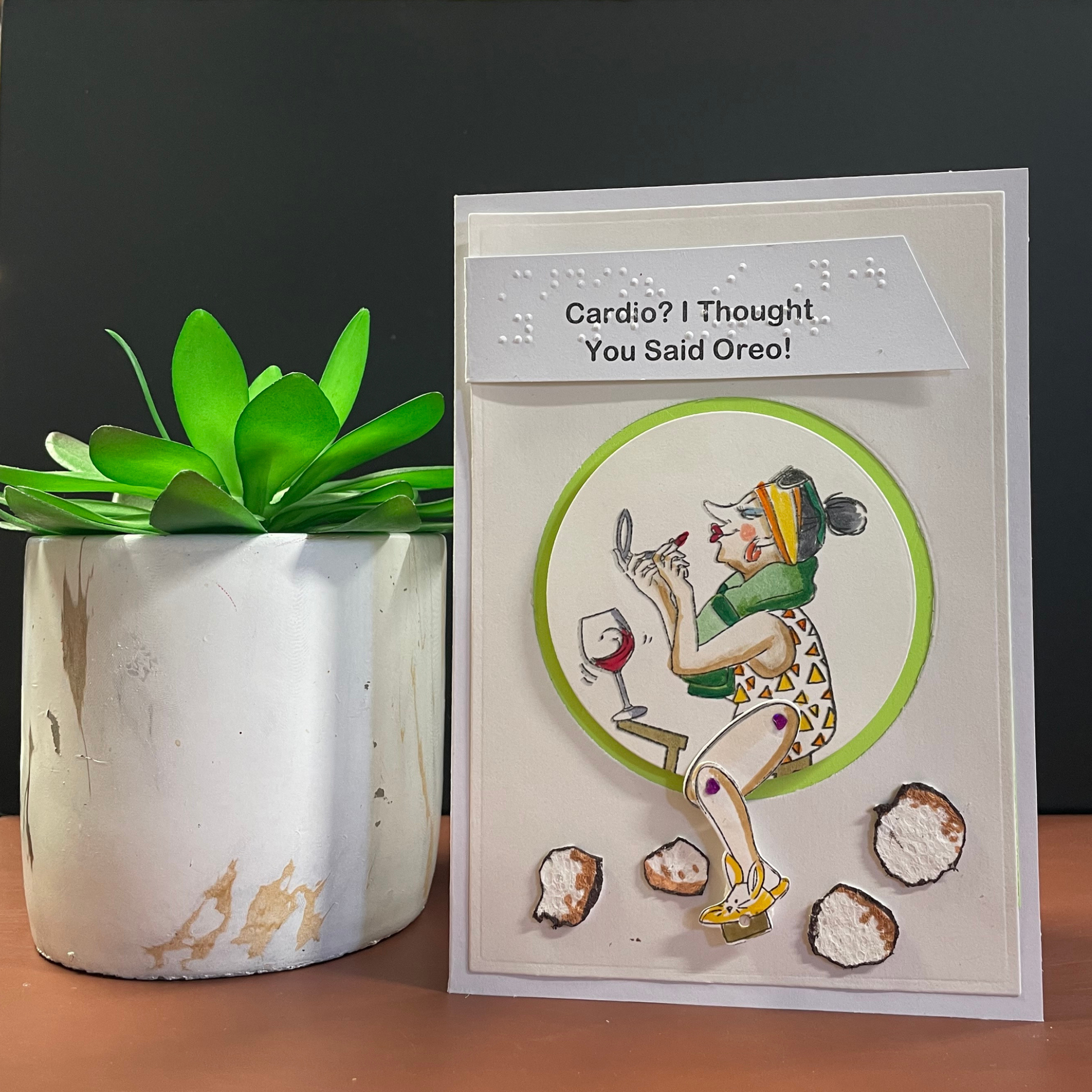 A handmade greeting card with a humorous caption reading 'Cardio? I Thought You Said Oreo!' displayed next to a potted green plant on a wooden surface against a dark gray background.