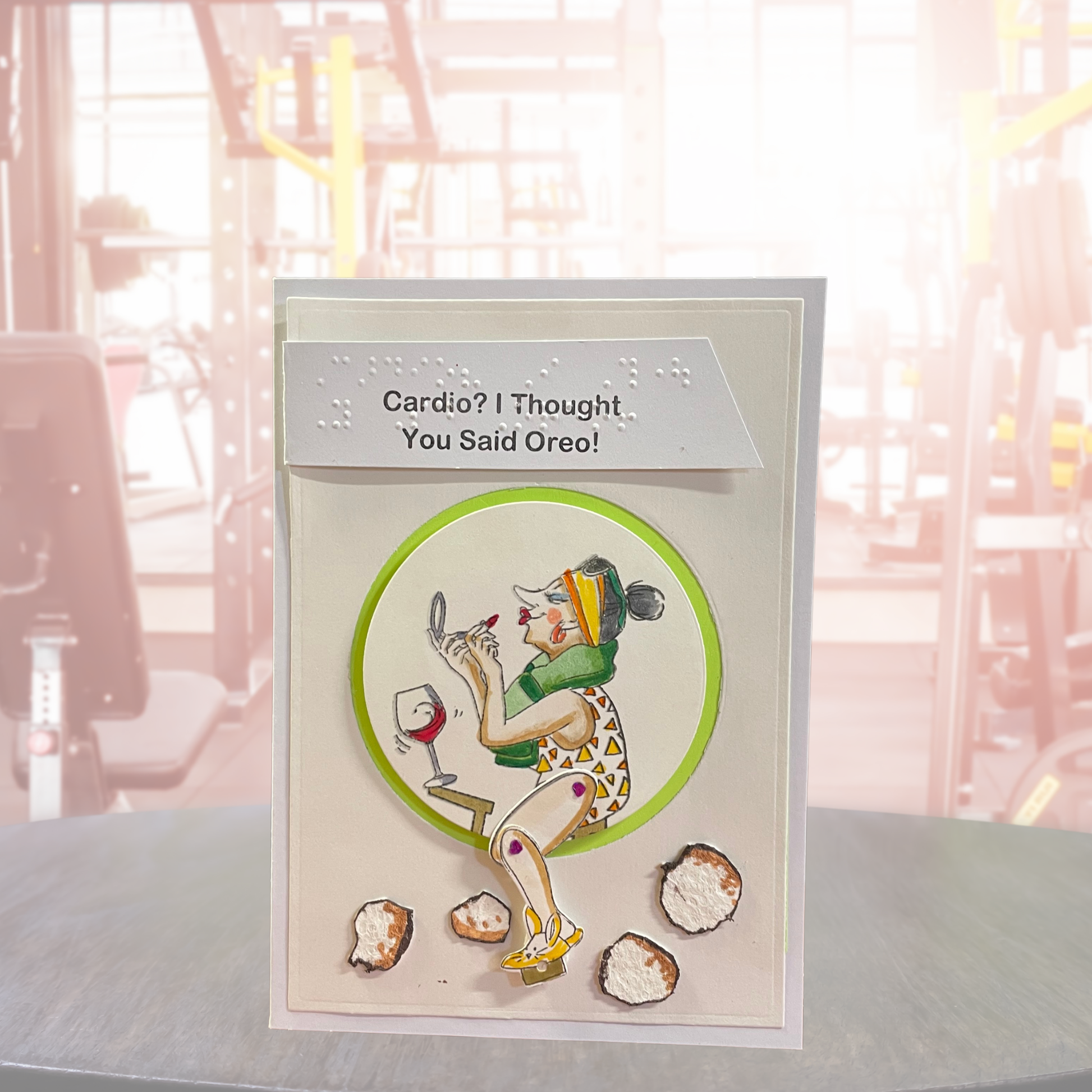A handmade greeting card featuring a retro-style illustration of a woman exercising with a wine glass and cookies, with the text 'Cardio? I Thought You Said Oreo!' at the top. The card is displayed on a table with a blurred gym background.