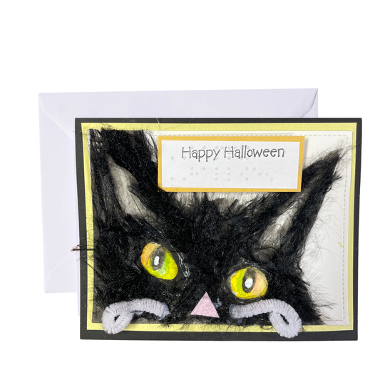 A Halloween greeting card featuring a black cat made from textured black yarn, with yellow eyes, a pink nose, and white pipe cleaners for whiskers, shown with a white envelope behind it on a white background.