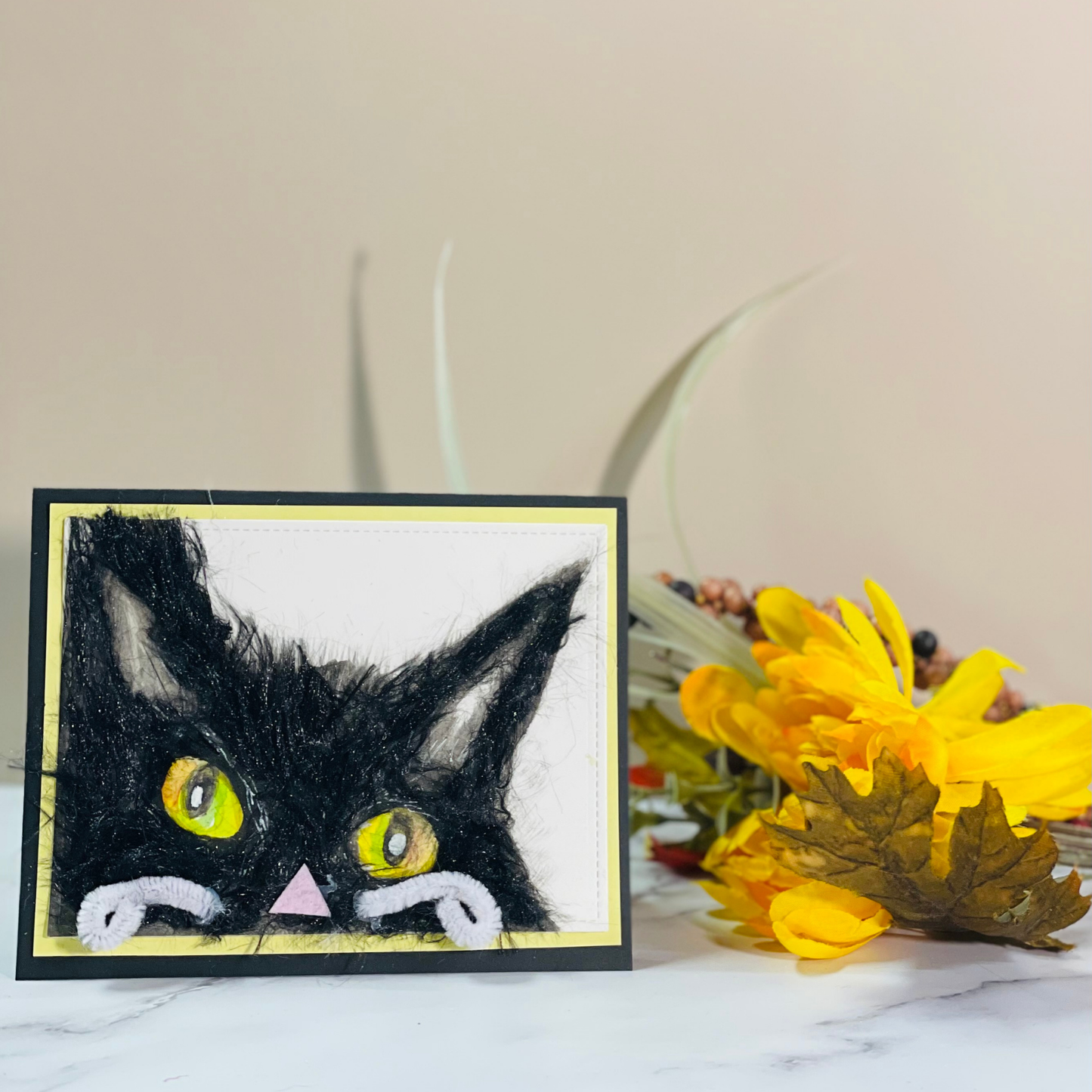 A handmade black cat card with a textured, fuzzy black cat face, yellow eyes, and a small pink nose, displayed on a marble surface next to yellow flowers against a beige background.