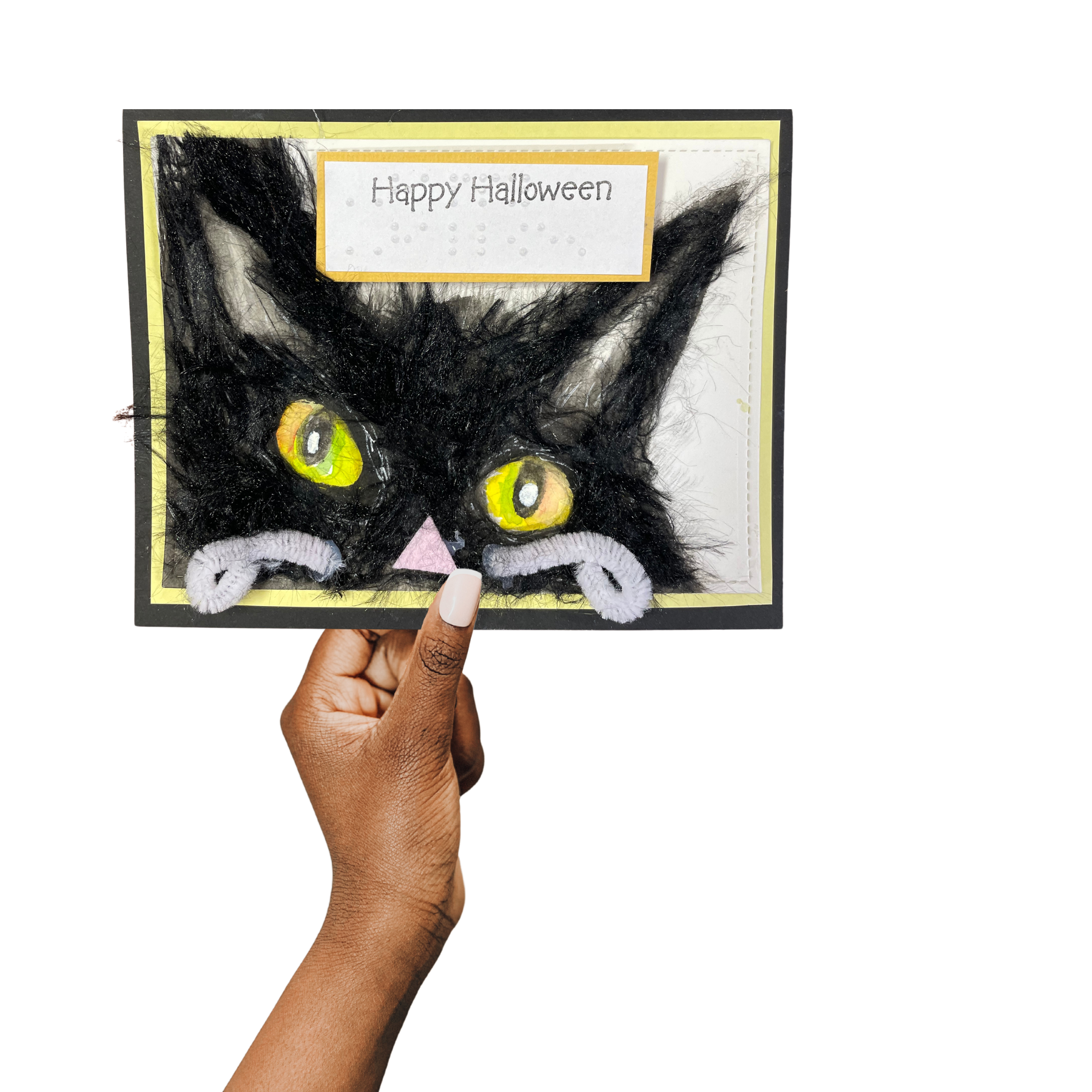 A handmade Halloween greeting card featuring a black cat with yellow eyes and a pink nose, held up by a hand against a plain white background. The card has 'Happy Halloween' written on a label at the top.