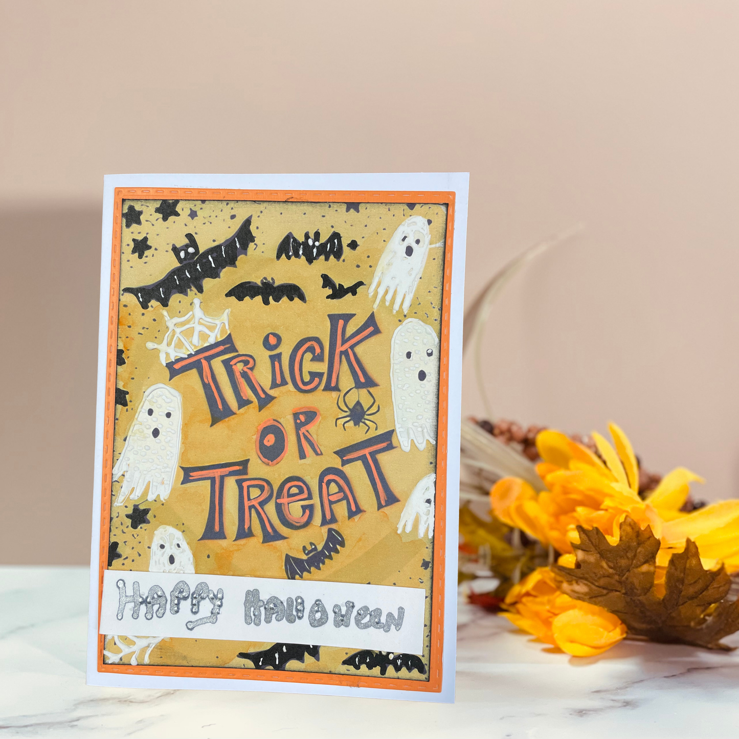 A handmade Halloween greeting card with 'Trick or Treat' and 'Happy Halloween' text, styled on a marble surface next to autumn leaves against a beige background.