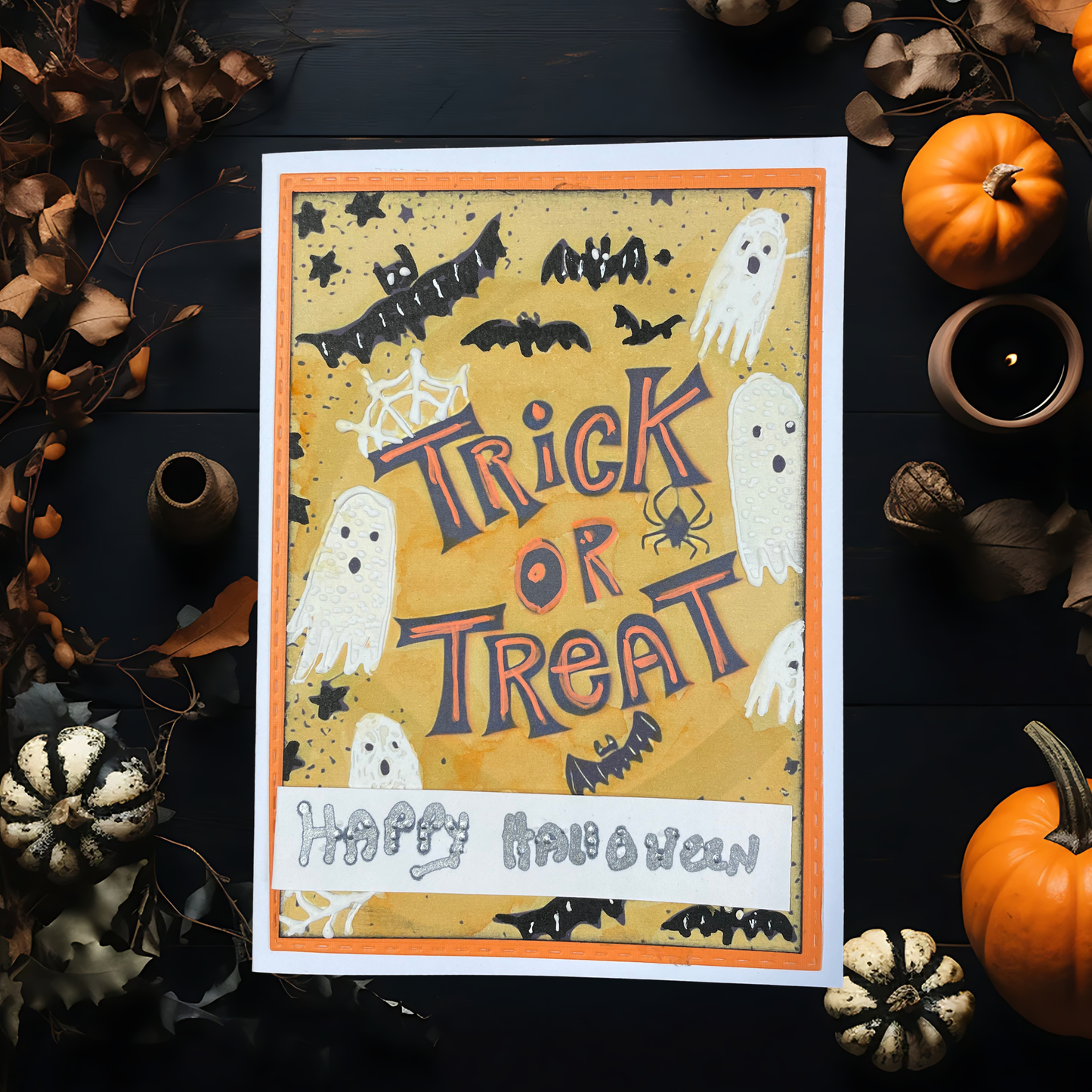 A Halloween greeting card with a whimsical 'Trick or Treat' design, styled in a flat-lay arrangement with miniature pumpkins, dried leaves, and a candle on a dark wooden surface.