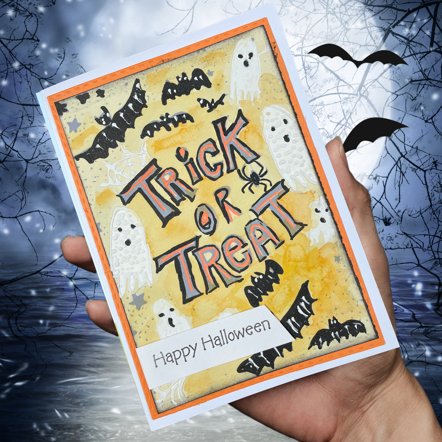 A hand holds a Halloween greeting card featuring the words 'Trick or Treat' in stylized lettering, with ghost, bat, spider, and star illustrations on a yellow background. The card is shown against a spooky night scene with bare tree branches and a full moon.
