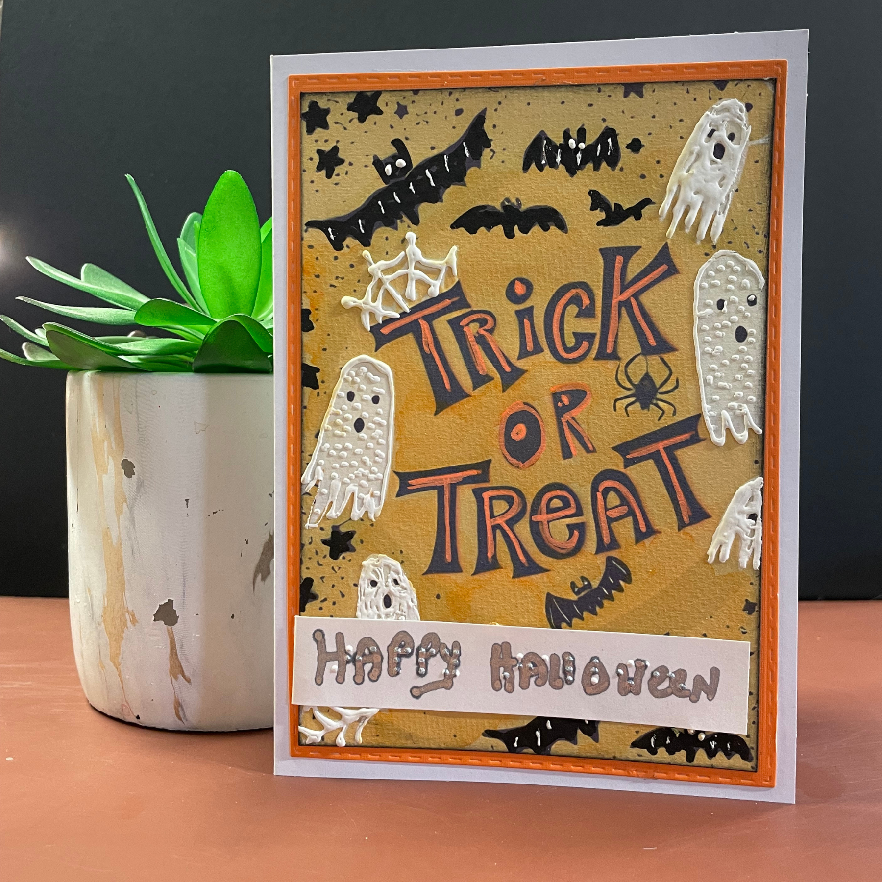A handmade Halloween greeting card with 'Trick or Treat' and 'Happy Halloween' text, decorated with white ghost cutouts, black bats, and a spider on a tan textured background with an orange border. The card is displayed standing upright on a wooden surface next to a potted succulent.