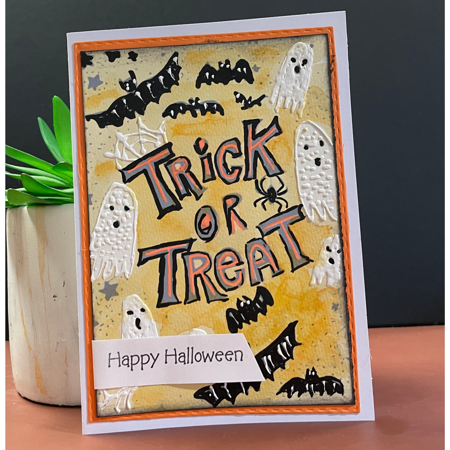 A handmade Halloween greeting card with 'Trick or Treat' in raised orange and black lettering, decorated with white ghosts, black bats, and a spider on a textured yellow background. A white banner at the bottom reads 'Happy Halloween'.