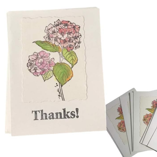A cream-colored greeting card featuring a watercolor-style illustration of pink hydrangea flowers with green leaves and the printed word 'Thanks!' below. A small stack of similar cards is fanned out on the right against a white background.