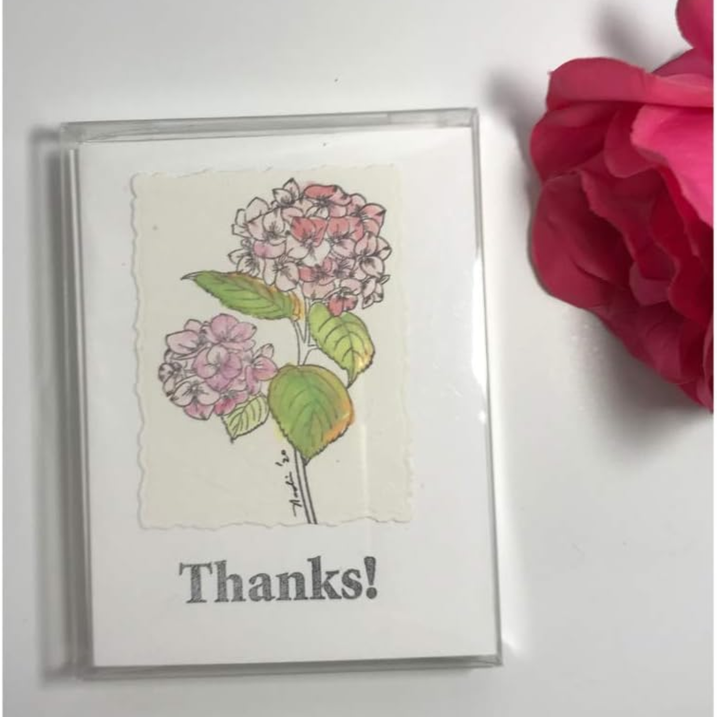 A square white card with a clear plastic sleeve, featuring an illustration of pink and white hydrangea flowers with green leaves and the text 'Thanks!' below. A large pink flower is partially visible on the right side against a white background.