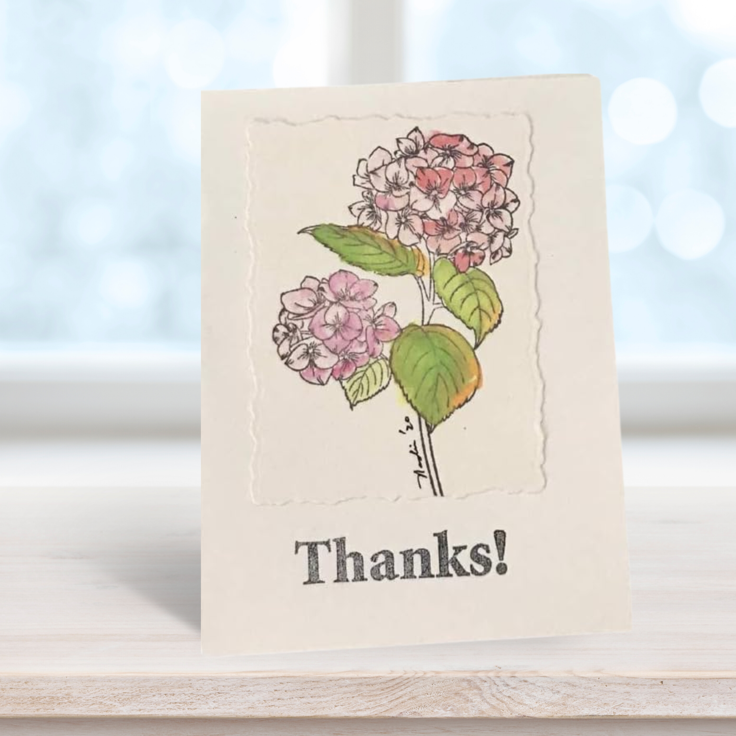 A handmade-style greeting card with a watercolor illustration of pink and white hydrangeas and the word 'Thanks!' printed below, standing on a light wooden surface with a blurred window background.