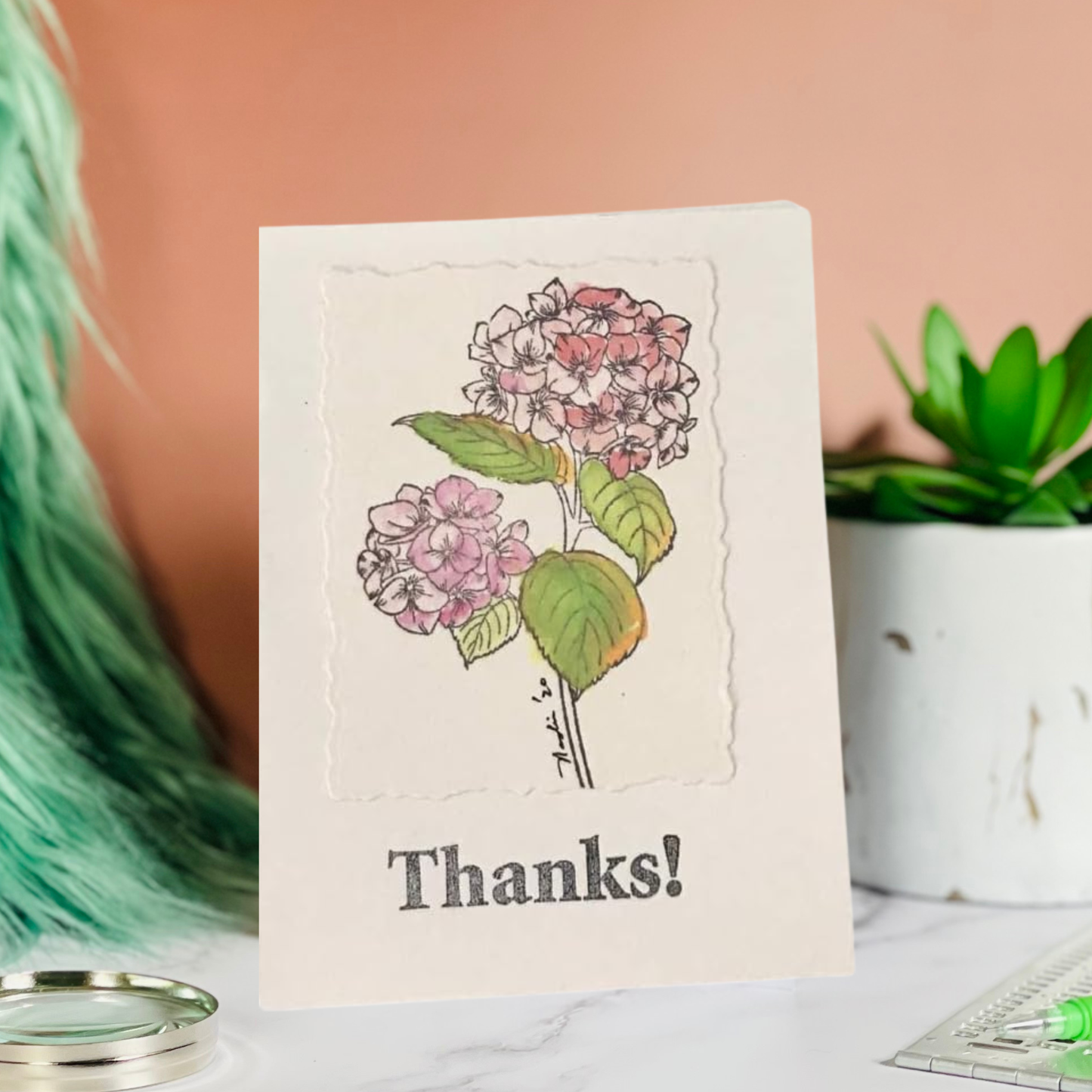 A handmade-style thank you card with a floral illustration of pink hydrangeas and the word 'Thanks!' printed at the bottom, displayed on a marble surface with a peach background and decorative props.