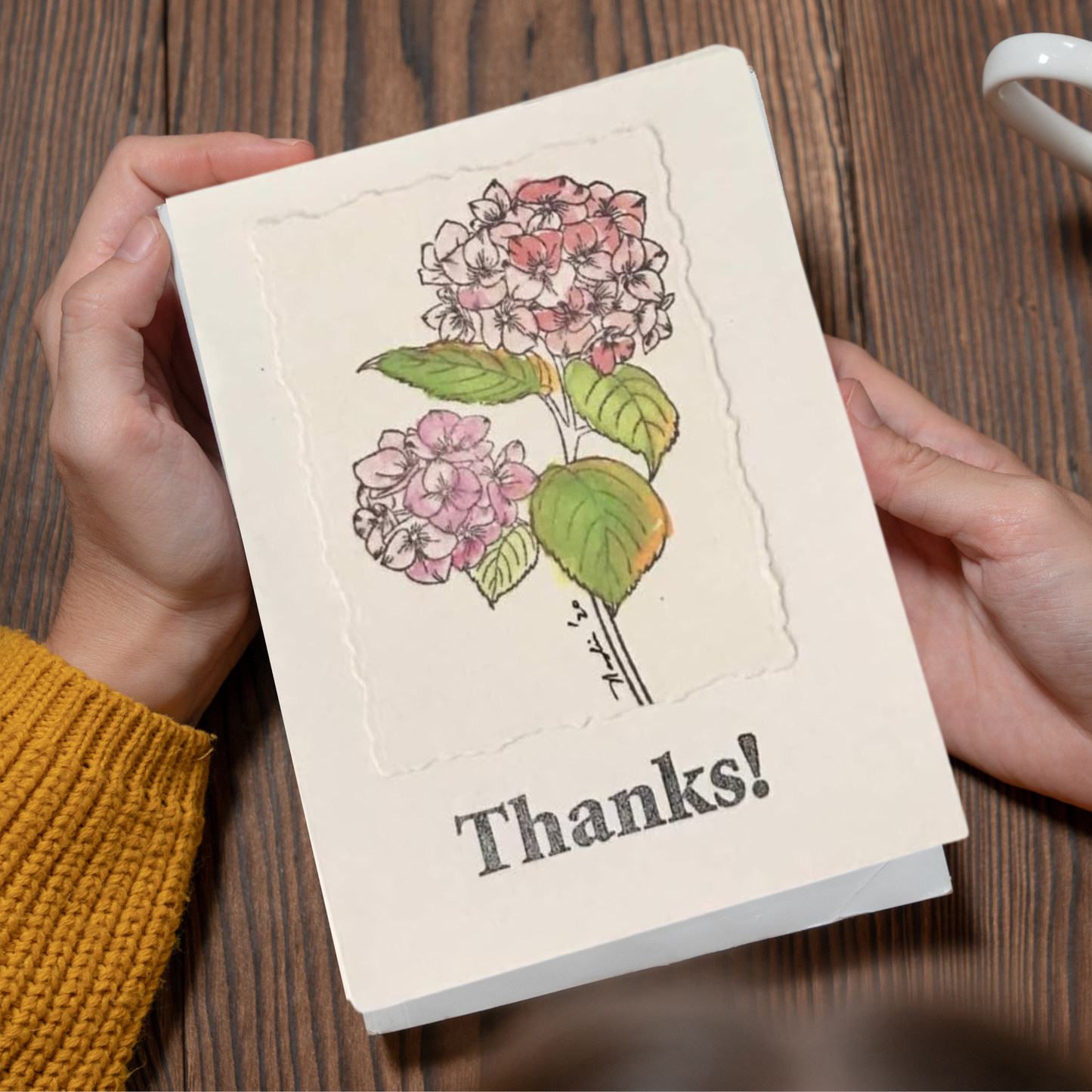 A person holds a cream-colored greeting card featuring an illustration of pink and white hydrangeas with green leaves and the word 'Thanks!' printed below. The card is held over a dark wood tabletop, with a person's hands and a yellow knit sleeve visible.