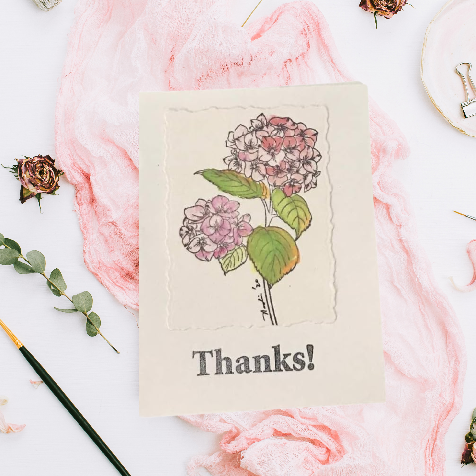 A handmade-style thank you card featuring an illustration of pink and white hydrangeas with green leaves and the text 'Thanks!' printed below, styled on a white surface with a pink fabric backdrop and dried floral props.