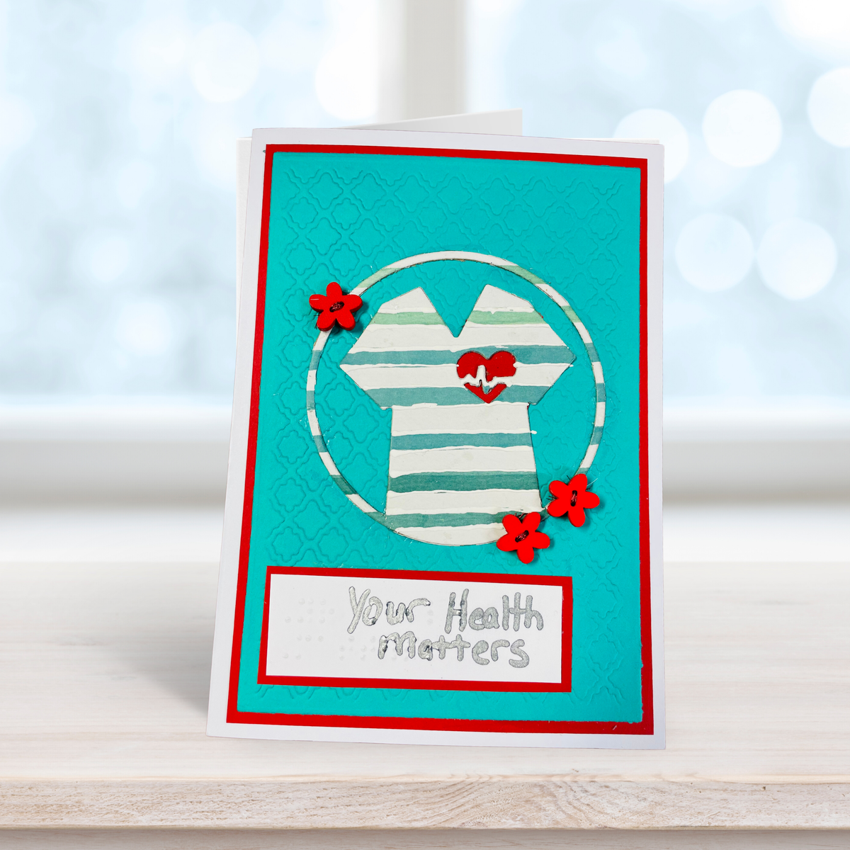 A handmade greeting card standing upright on a light wooden surface. The card features a turquoise background with a red border, a white and teal striped shirt graphic inside a circle, three red paper flowers, and a white banner at the bottom with the text 'Your Health Matters'.