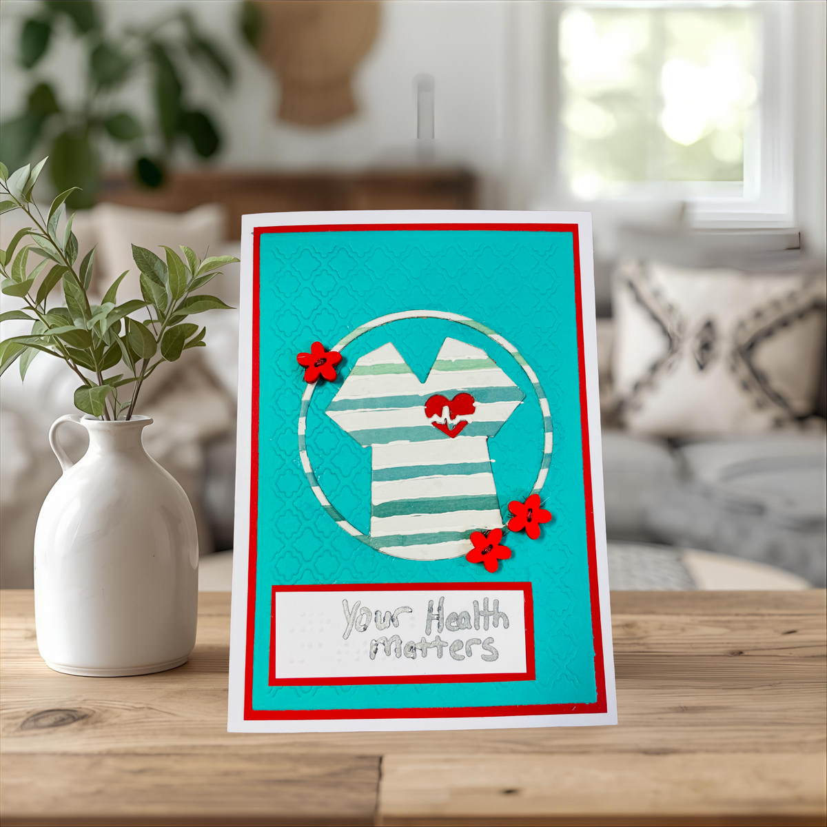 A handmade greeting card displayed on a wooden table next to a white ceramic vase with green leaves. The card features a turquoise background with a red border, a white and green striped shirt graphic with a red heart, and the text 'Your Health Matters'.