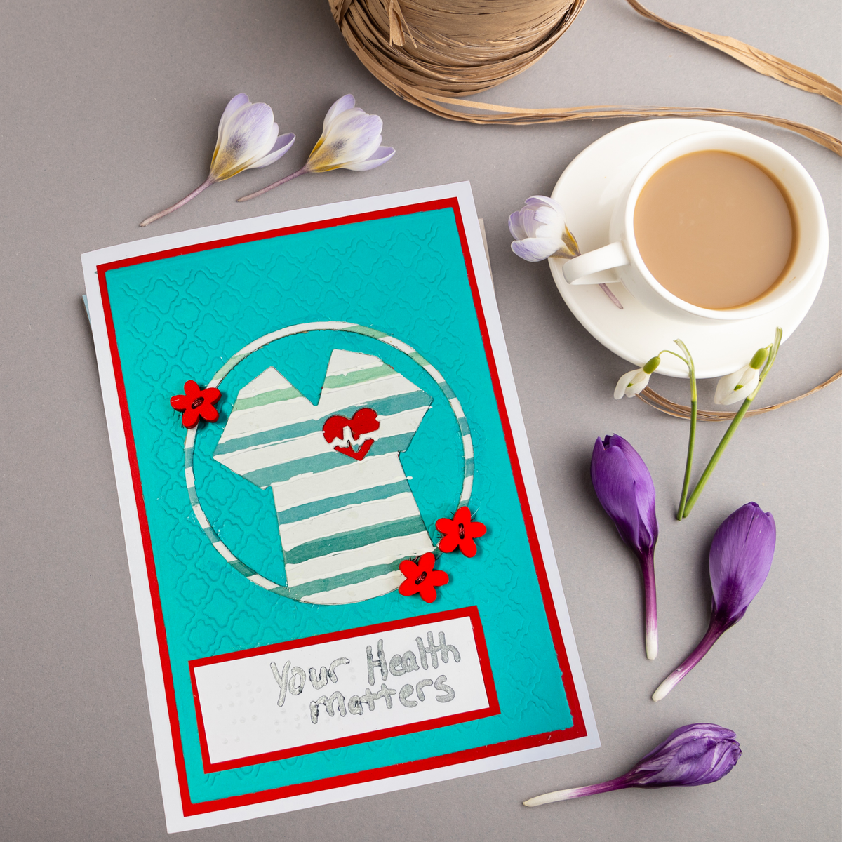 A handmade greeting card with a turquoise background, a white striped heart design, red flowers, and the text 'Your Health Matters', styled on a gray surface with a cup of coffee, twine, and purple flowers.
