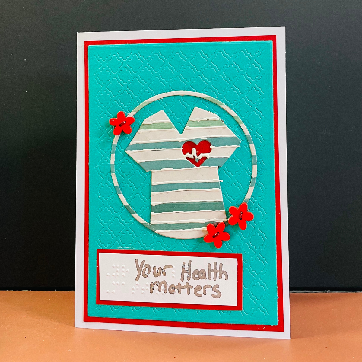 A handmade greeting card featuring a turquoise background with a white and green striped shirt layered inside a circular frame, accented with three red flowers. The bottom of the card has a white banner with the words 'Your Health Matters' in brown script, bordered in red.