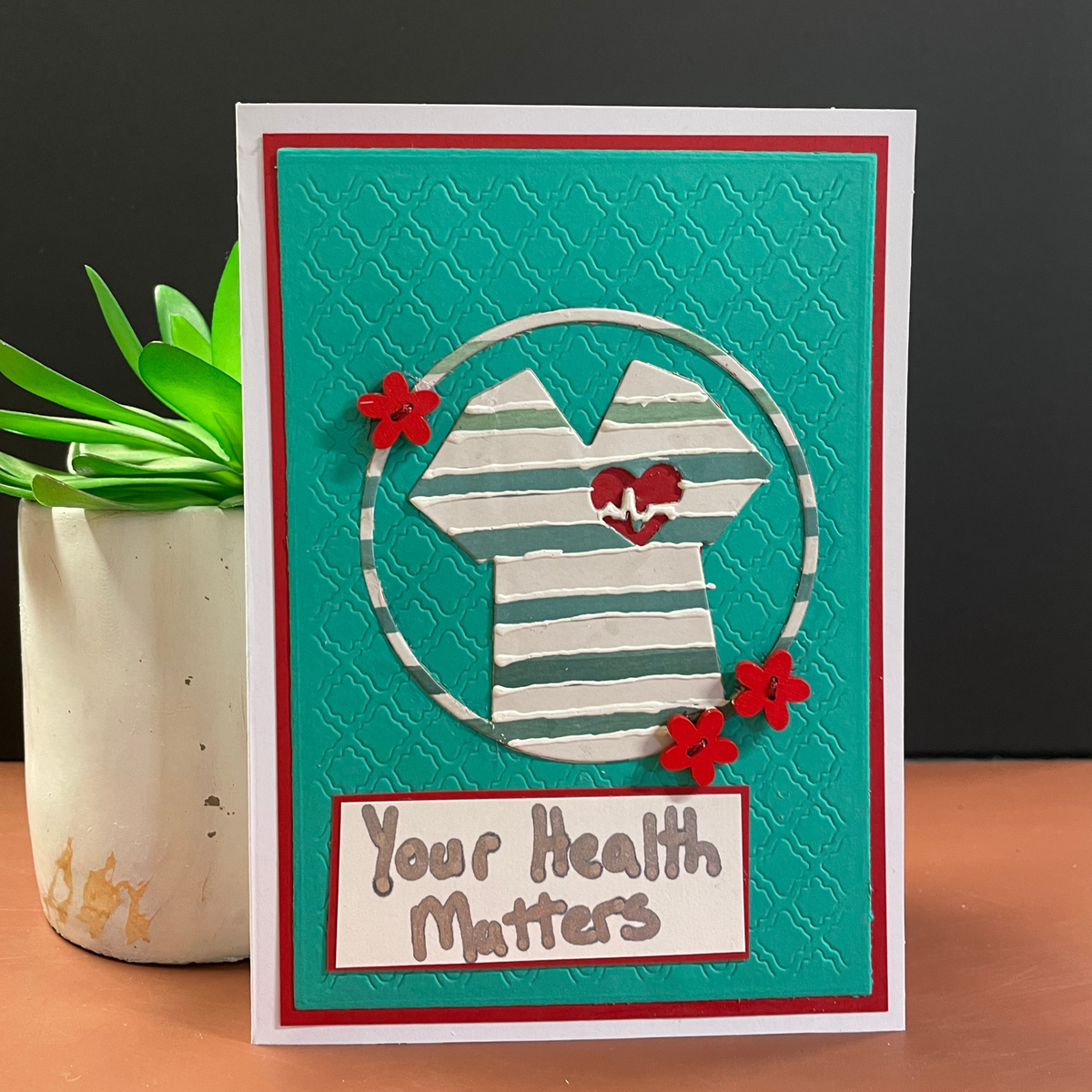 A handmade greeting card featuring a layered design with a teal textured background, a white and teal striped shirt cutout with red hearts, three red flowers, and a white banner reading 'Your Health Matters'. The card is displayed on a wooden surface next to a small potted succulent against a dark gray background.