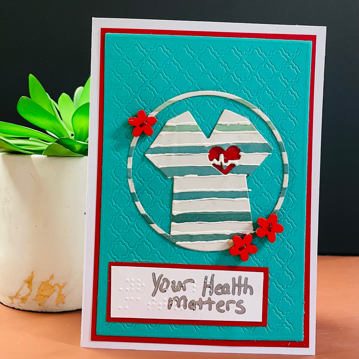 A handmade greeting card featuring a stylized striped shirt inside a circle on a turquoise textured background, with red flowers and the words 'Your Health Matters' at the bottom. The card is displayed on a wooden surface next to a potted plant against a dark background.