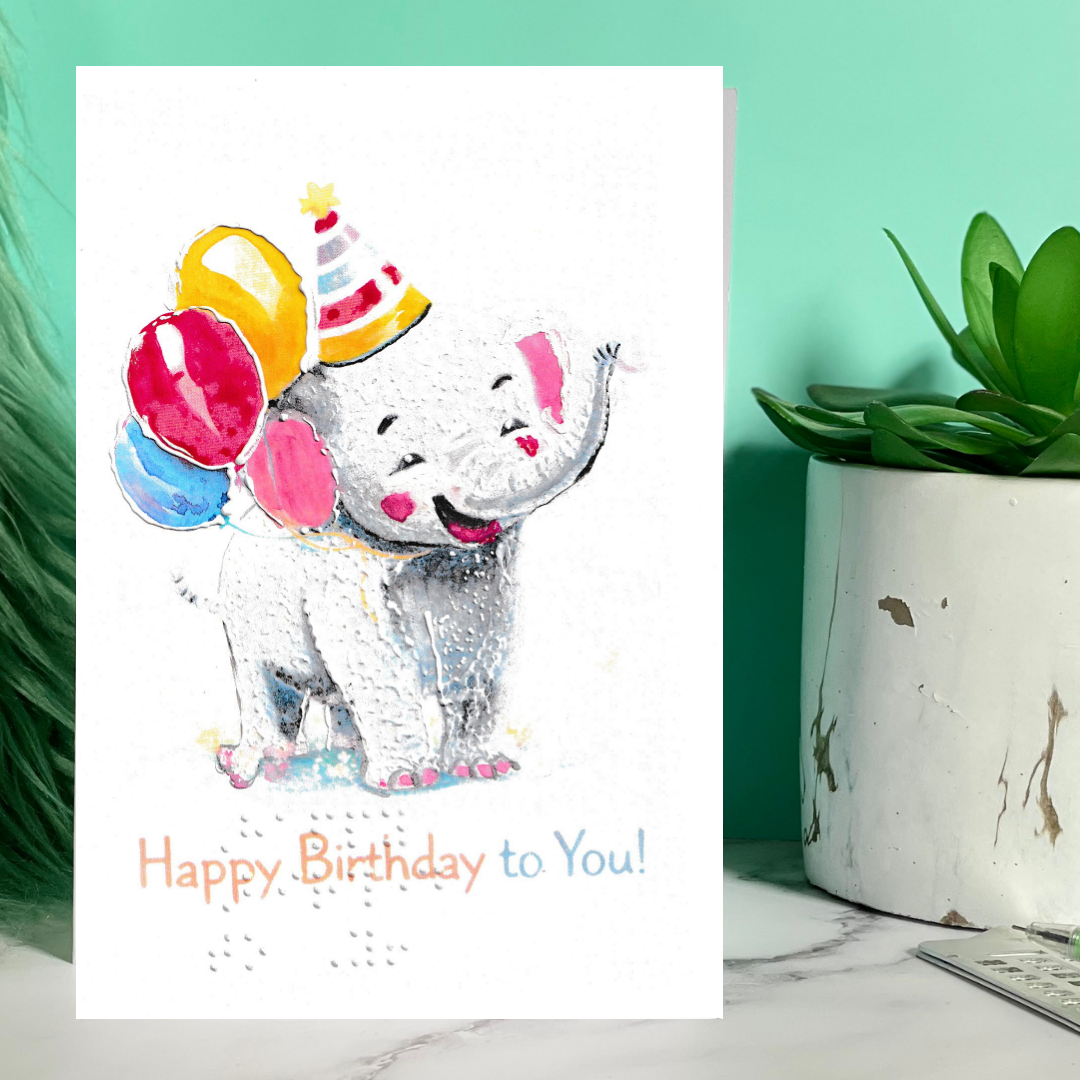 Birthday card with a cartoon elephant holding balloons on a marble surface with a plant.
