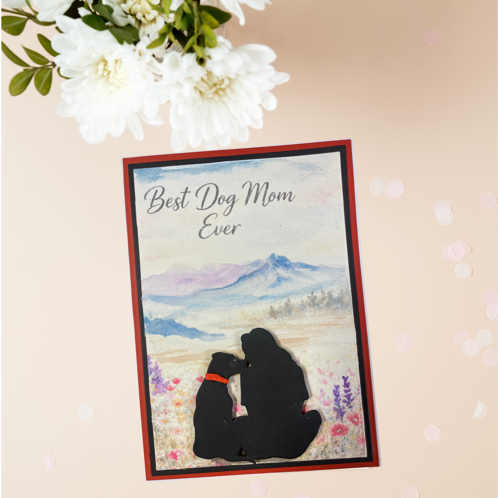  A beautiful black silhouette of a woman and her dog sitting side by side in a wildflower meadow, gazing at a soft watercolor mountain landscape — framed by a bold red and black border. 
