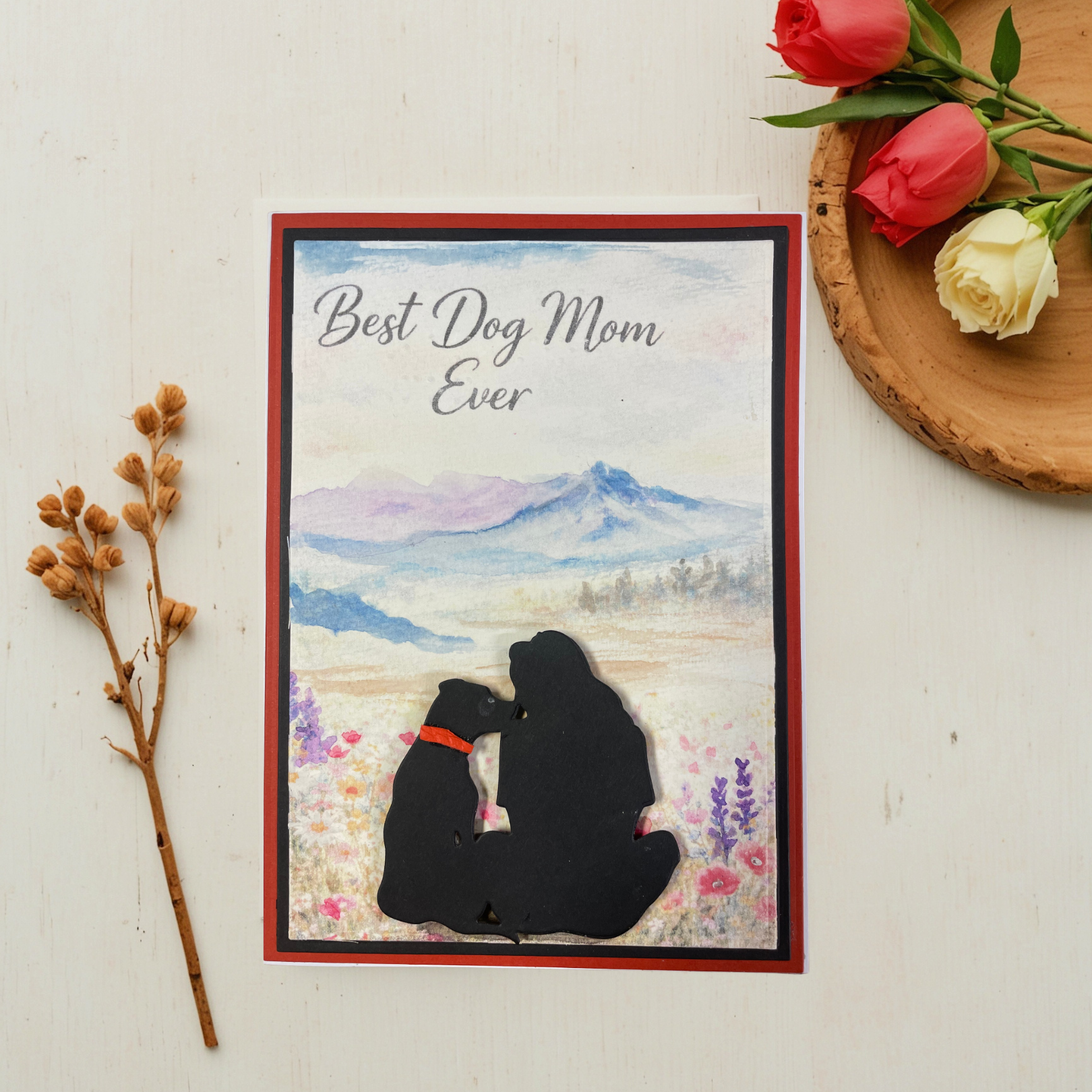 Greeting card with 'Best Dog Mom Ever' text and dog and person silhouette, placed on a surface with flowers and branches.