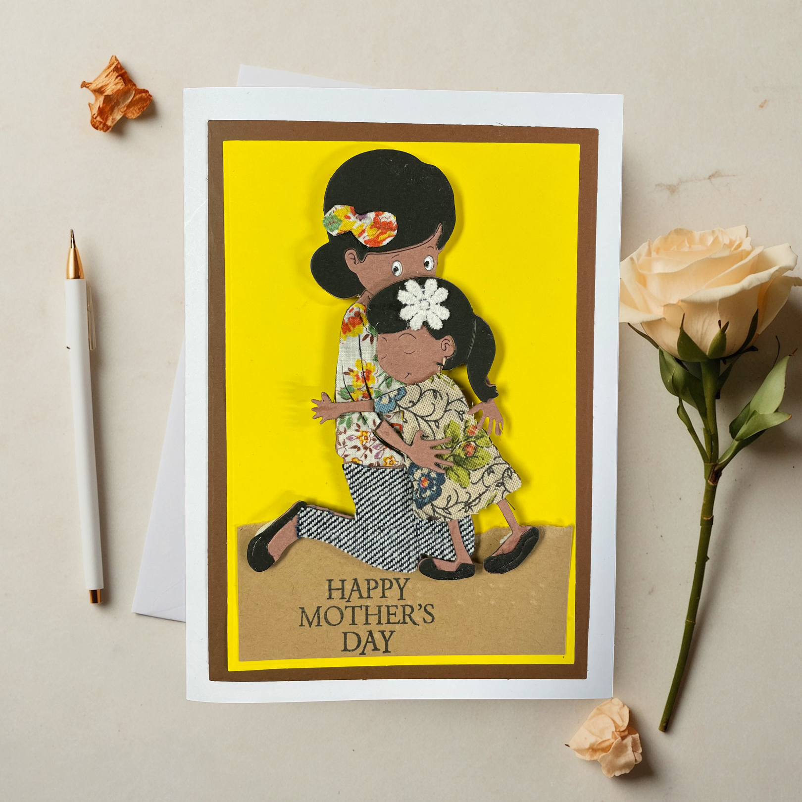 Greeting card with tactile image of a mother and daughter on a yellow background, with flowers and a pen on the side.