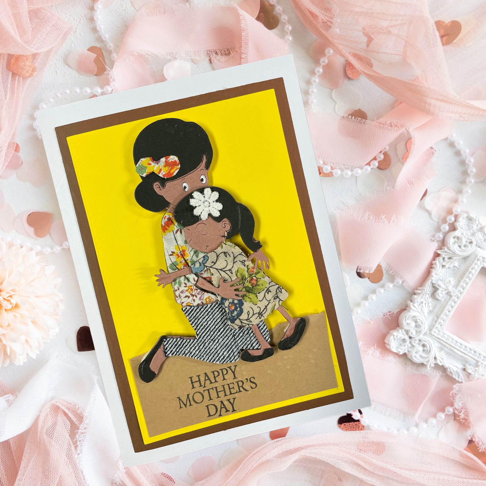 Mother's Day card with a tactile image of a mother and child on a yellow background, surrounded by pink flowers and ribbons.