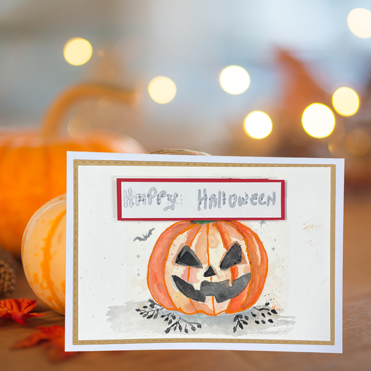 A handmade Halloween greeting card featuring a watercolor illustration of a carved orange pumpkin with a smiling face and the words 'Happy Halloween' in gray script inside a red-bordered box. The card is displayed on a wooden surface with blurred pumpkins and warm bokeh lights in the background.