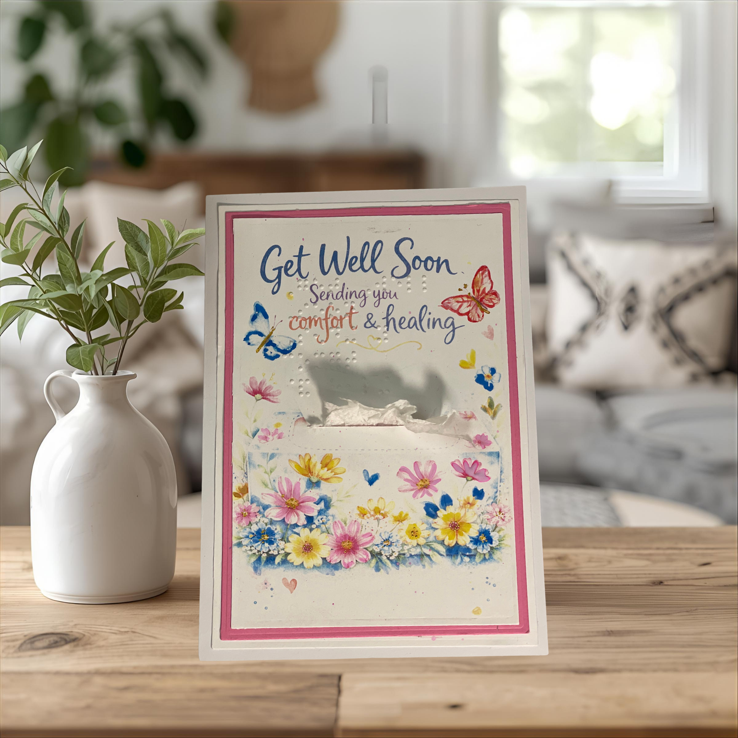 Get Well Soon card with flowers and butterflies on a wooden table with a vase and plant in the background.