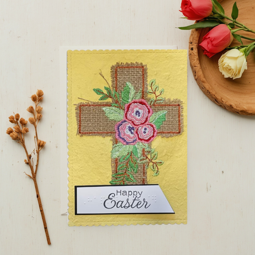 Easter card with floral cross design and 'Happy Easter' text, placed on a light surface with flowers and branches.