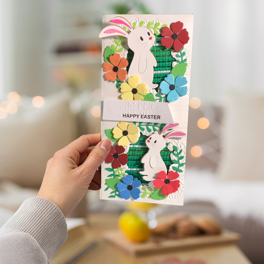 Hand holding a colorful Easter card with bunnies and flowers against a blurred indoor background.