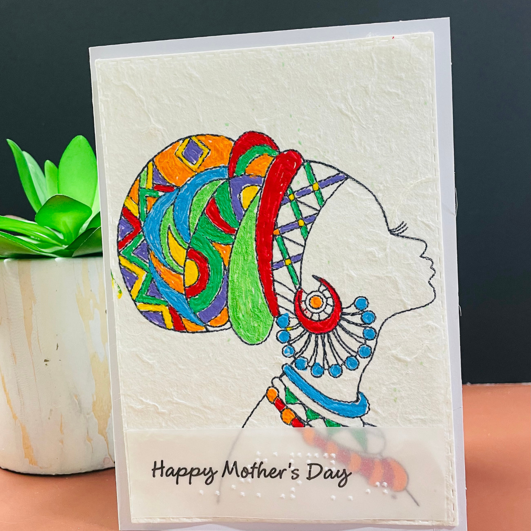 Mother's Day card features a hand-drawn profile silhouette of a woman adorned with a vivid geometric head wrap in orange, red, green, blue, and purple, paired with layered statement jewelry in matching jewel tone 'Happy Mother's Day' text, placed on a surface with a plant in the background.