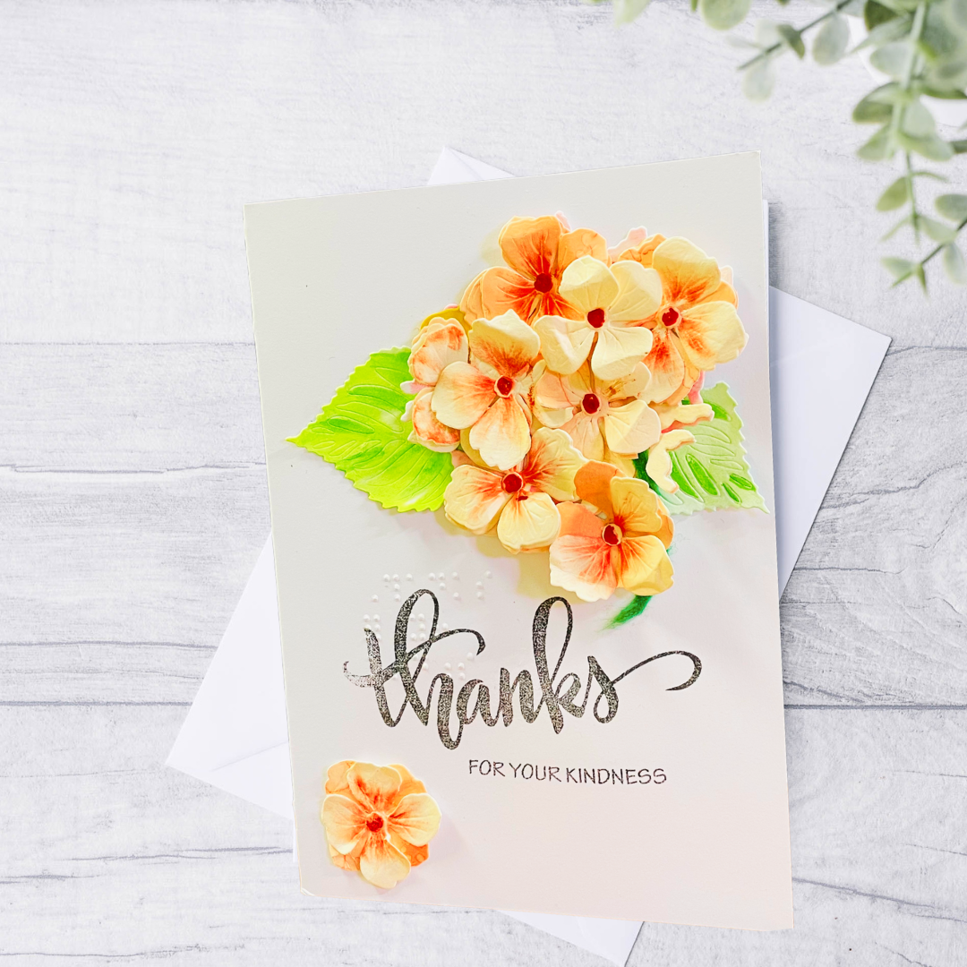 A handmade-style thank you card with Peach Hydrangea Bouquet green leaves, and metallic silver 'thanks' script, displayed on a white envelope over a light gray wood background.