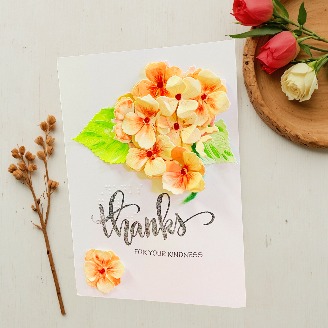 A handmade greeting card with a 3D floral design, featuring Peach Hydrangea Bouquet with green leaves and the text 'thanks FOR YOUR KINDNESS' in black. The card is styled on a white wooden surface with dried stems and a wooden tray holding red and white roses.
