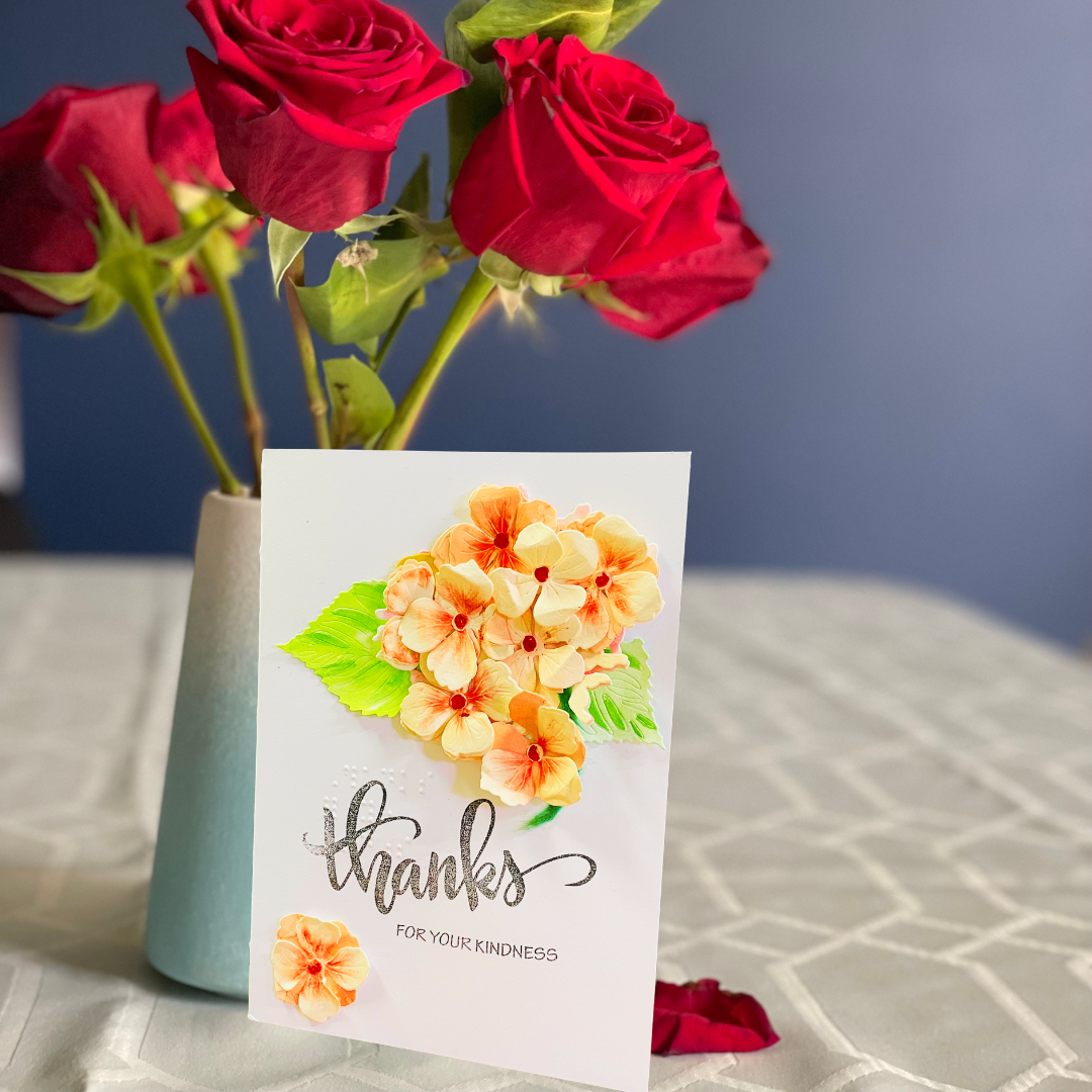 A white greeting card with a watercolor Peach Hydrangea Bouquet and the words 'thanks FOR YOUR KINDNESS' in gray script, styled in front of red roses in a light blue vase on a white textured tablecloth against a blue wall.