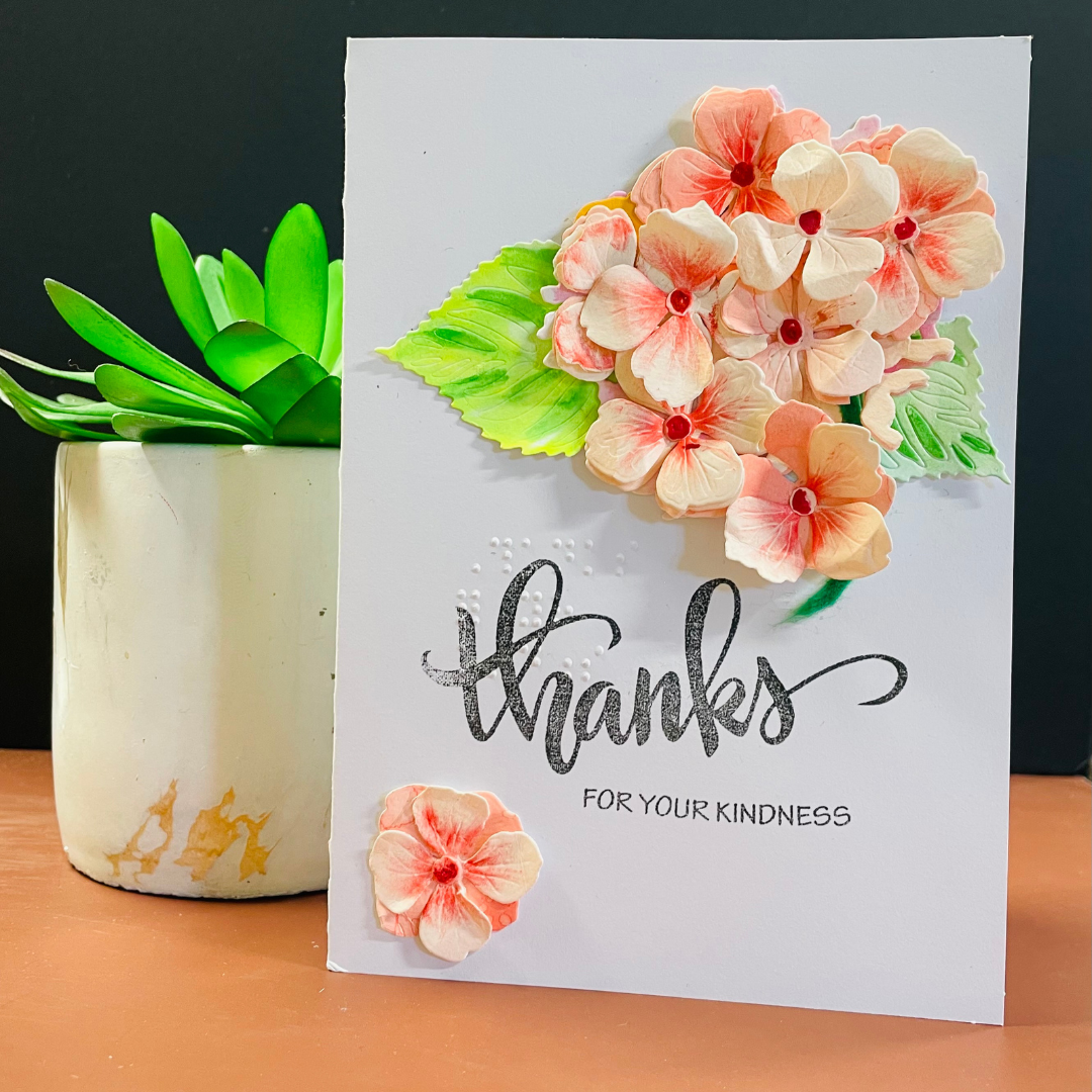 A handmade white greeting card with 3D Peach Hydrangea Bouquet, green leaves, and the text 'thanks FOR YOUR KINDNESS' in black script and block lettering. The card is displayed on a wooden surface next to a potted green succulent against a dark background.