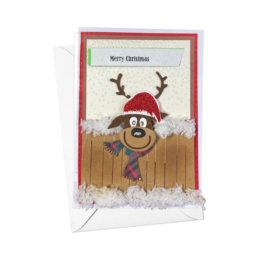 A handmade Christmas greeting card featuring a cartoon reindeer wearing a red Santa hat and plaid scarf, set against a snowy white background with a wooden fence. The card is shown with its white envelope behind it on a clean white background.