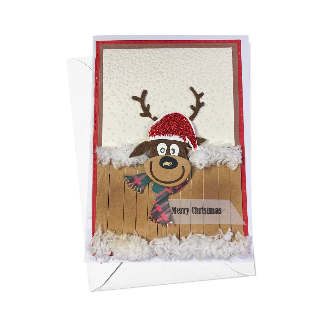 A handmade Christmas greeting card featuring a cartoon reindeer wearing a red Santa hat and plaid scarf, with a wooden fence and white fluffy trim. The card is shown with its white envelope against a plain white background.