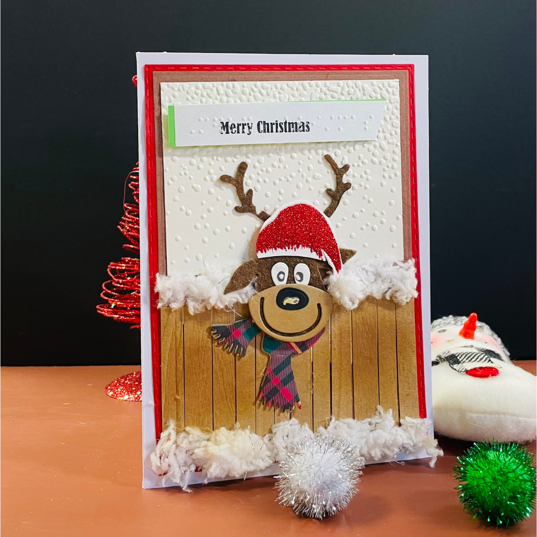 A handmade Christmas greeting card featuring a cartoon reindeer wearing a red Santa hat and plaid scarf, peeking over a wooden fence with white fluffy trim. The card is displayed on a tabletop with holiday-themed props against a dark background.
