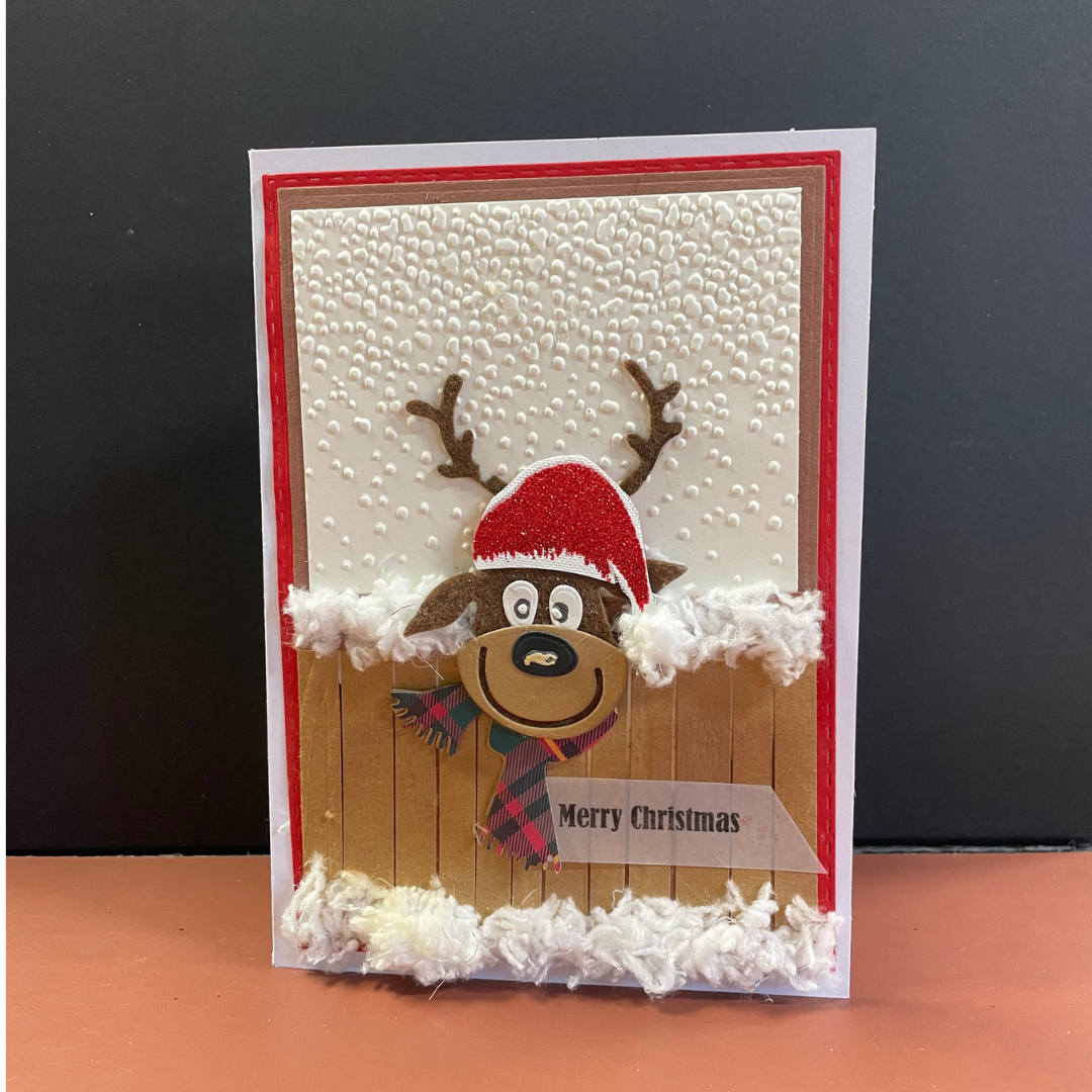 A handmade Christmas greeting card featuring a cartoon reindeer wearing a red Santa hat and plaid scarf in front of a faux wood fence, with white faux snow details and a 'Merry Christmas' banner, set against a dark gray background.