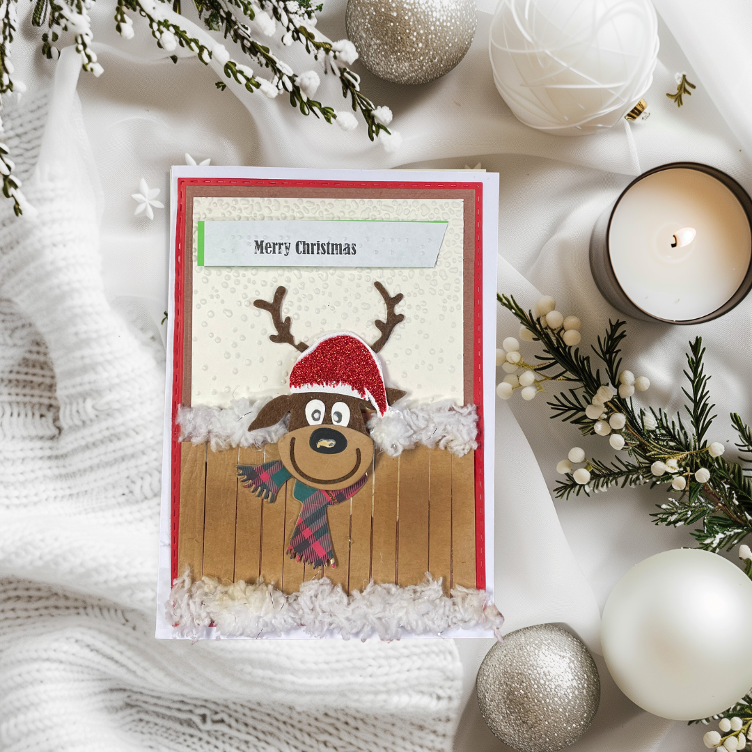 A handmade Christmas greeting card featuring a cartoon reindeer wearing a red Santa hat and plaid scarf, set against a snowy winter scene. The card is styled in a flat-lay arrangement with a white knit blanket, pine branches, silver and white ornaments, and a lit candle.