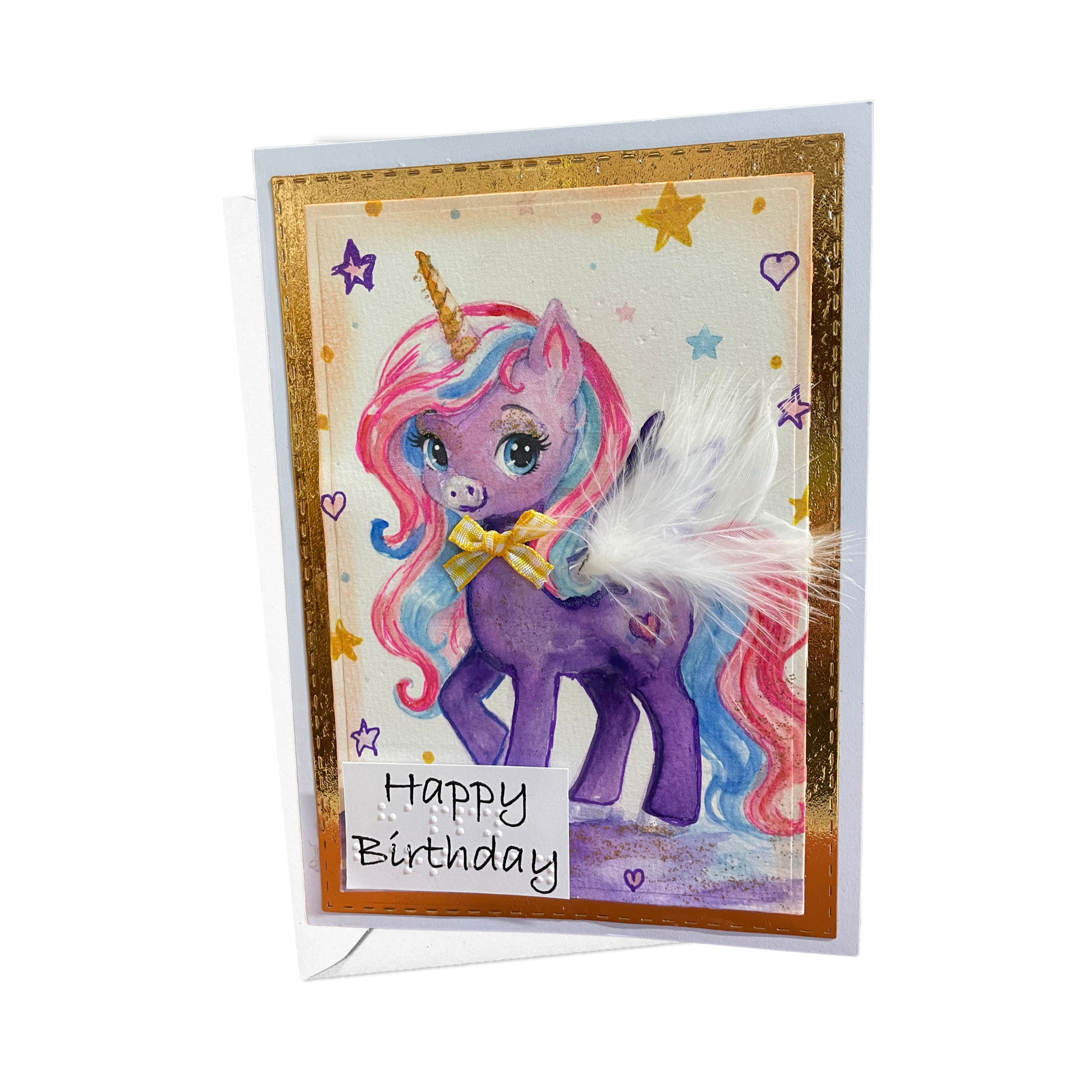 A handmade birthday card featuring a purple unicorn with a pink and blue mane, gold horn, and a white feather accent. The card has a gold glitter border, 'Happy Birthday' written in print and braille on a white label, and is displayed on a white background.