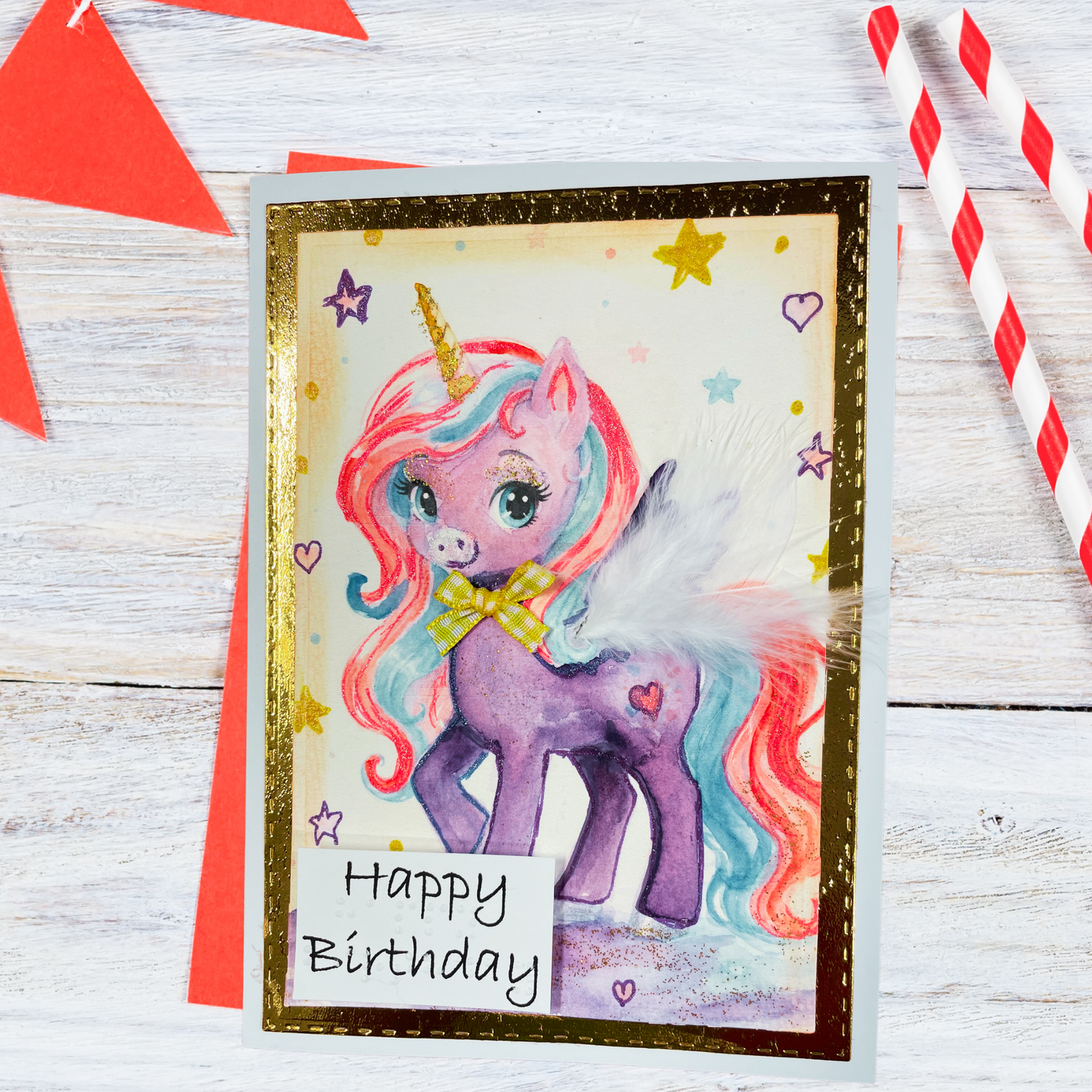 A handmade unicorn birthday card featuring a purple pony with a pink, blue, and yellow mane, set against a glittery gold border. The card is displayed on a white wooden surface with red paper decorations and striped straws.