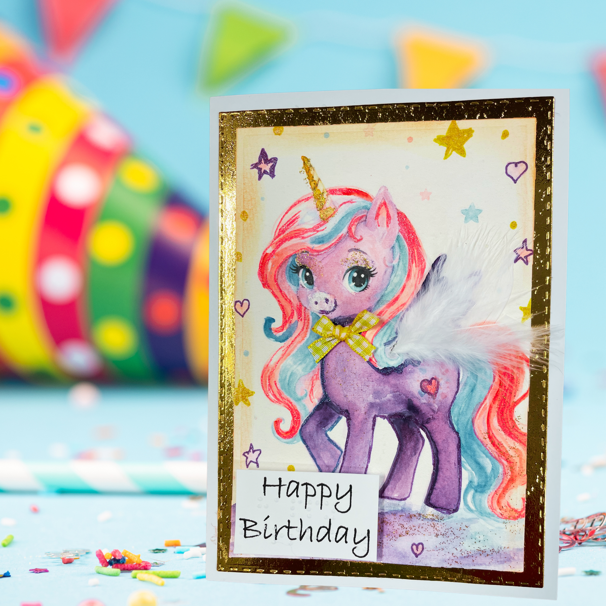 A handmade unicorn birthday card with a purple pony, colorful hair, and a gold glitter border, displayed on a light blue surface with a blurred party hat in the background.
