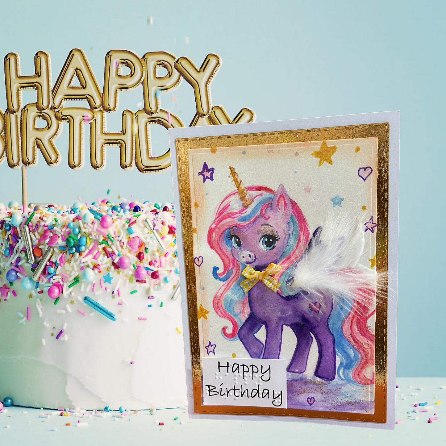 A handmade unicorn birthday card with a purple pony, pastel rainbow mane, and gold foil border, displayed next to a decorated cake with a gold 'HAPPY BIRTHDAY' topper against a light blue background.