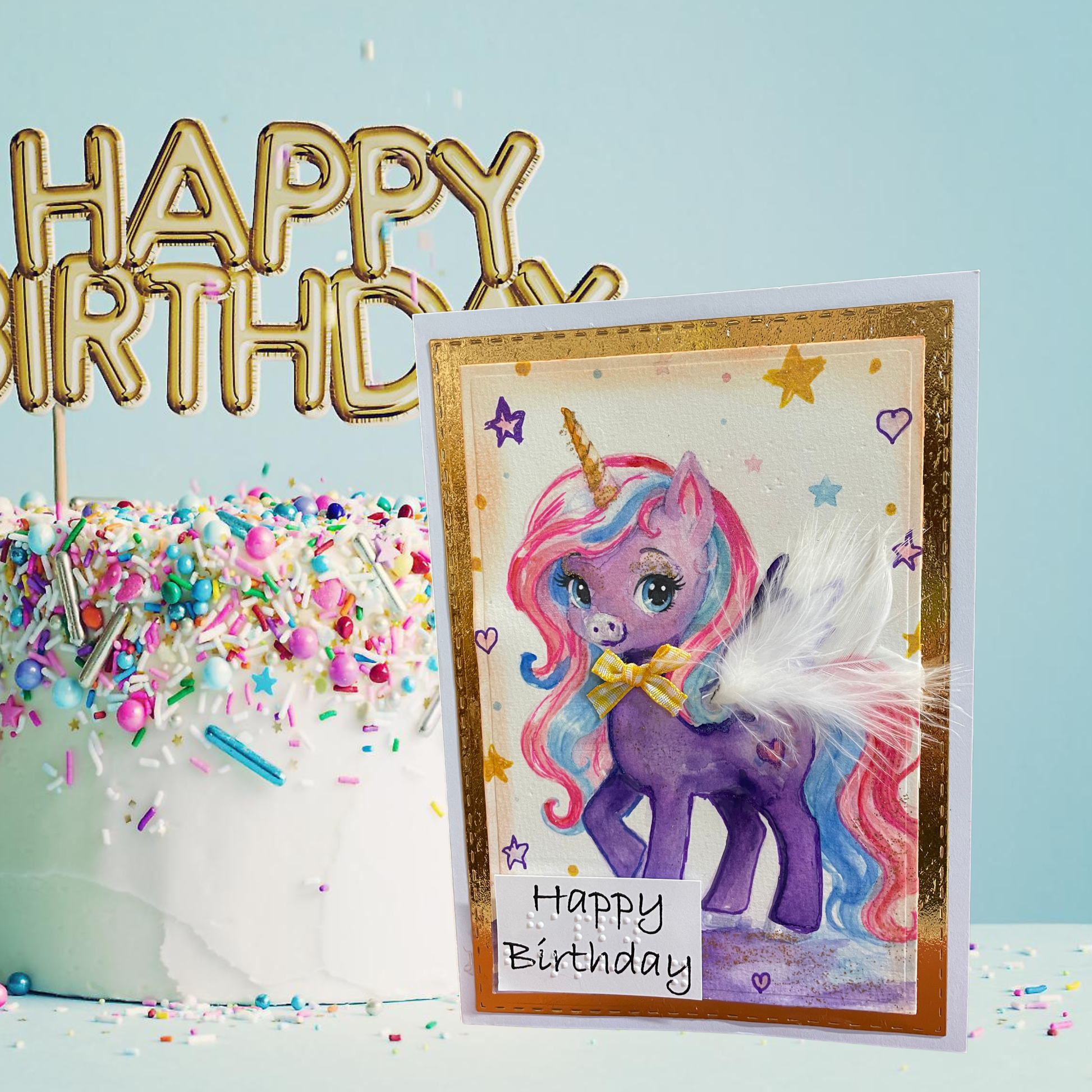 A handmade unicorn birthday card with a purple pony, pastel rainbow mane, and gold foil border, displayed next to a decorated cake with a gold 'HAPPY BIRTHDAY' topper against a light blue background.
