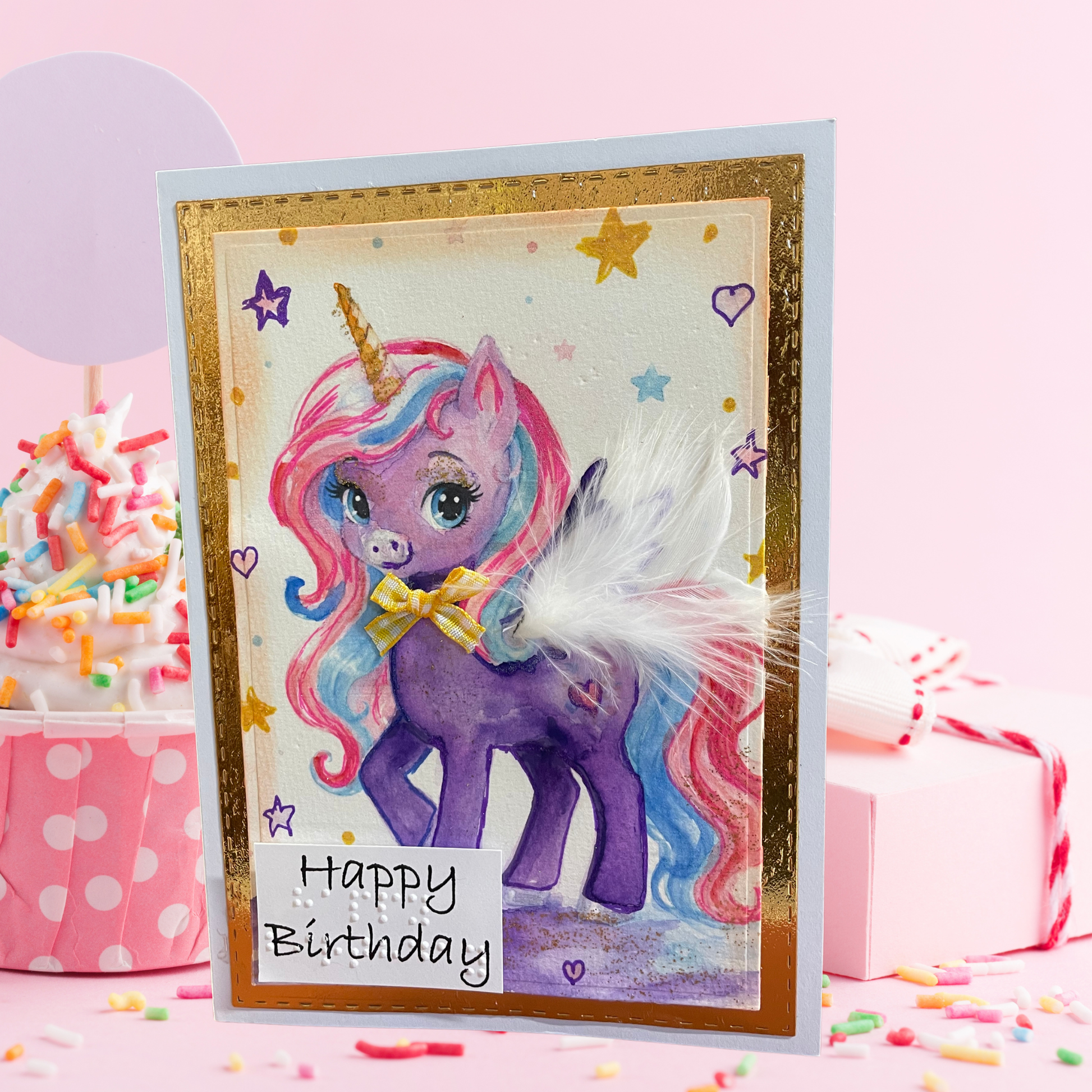 A handmade unicorn birthday card with a purple unicorn illustration, pastel rainbow mane and tail, gold glitter border, and a white feather accent, displayed on a pink background with cupcakes and a gift box.